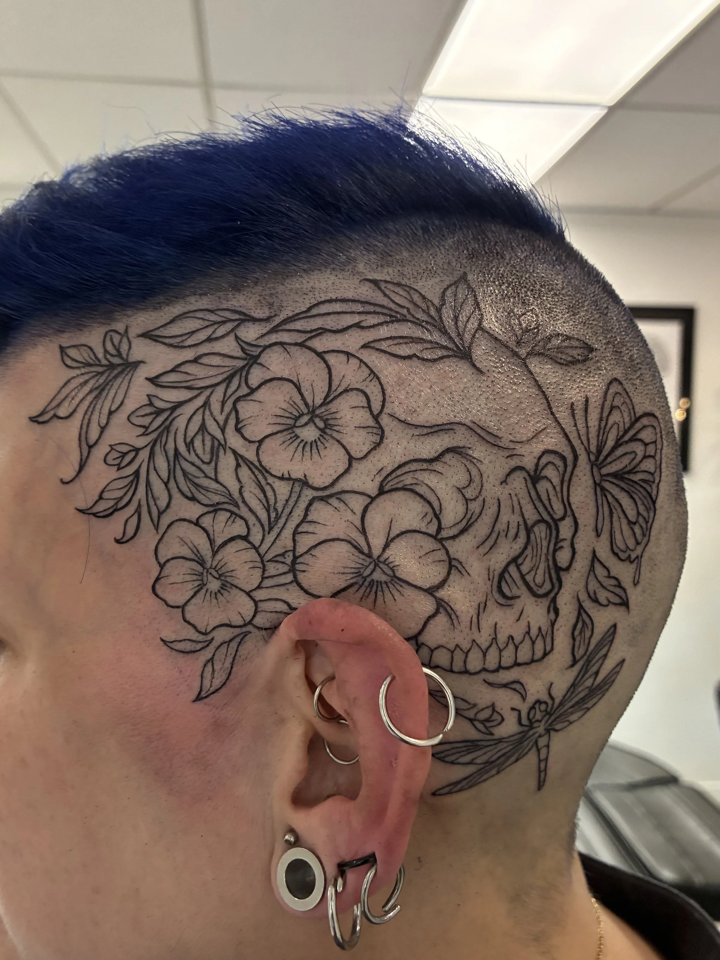 Close-up photograph of a person's head featuring a tattoo of a skull with flowers and leaves, with bright blue hair on top and multiple earrings in the ear.