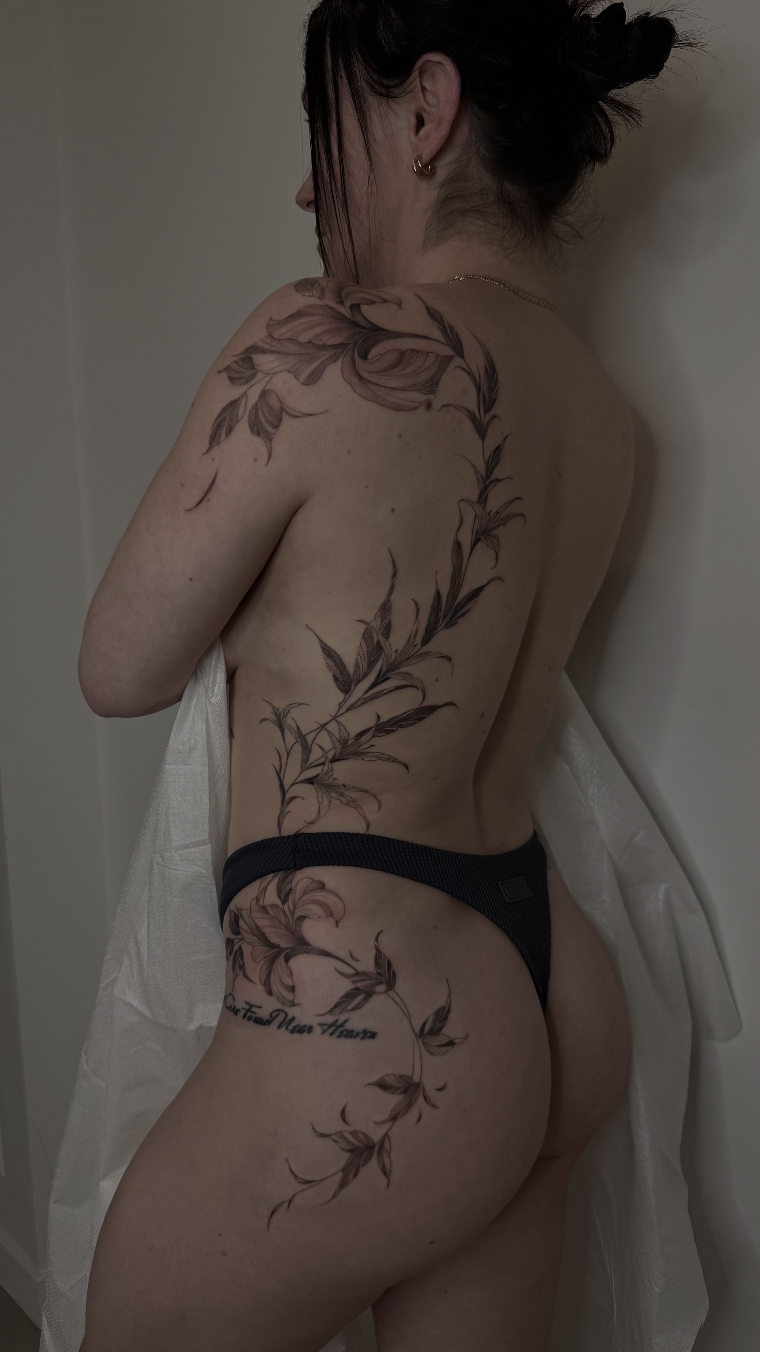 A woman with dark hair styled in buns or ponytails, wearing gold hoop earrings, a gold chain necklace, and black underwear. She has a large floral tattoo with leaves and flowers running from her shoulder down her side and thigh. She is leaning agains