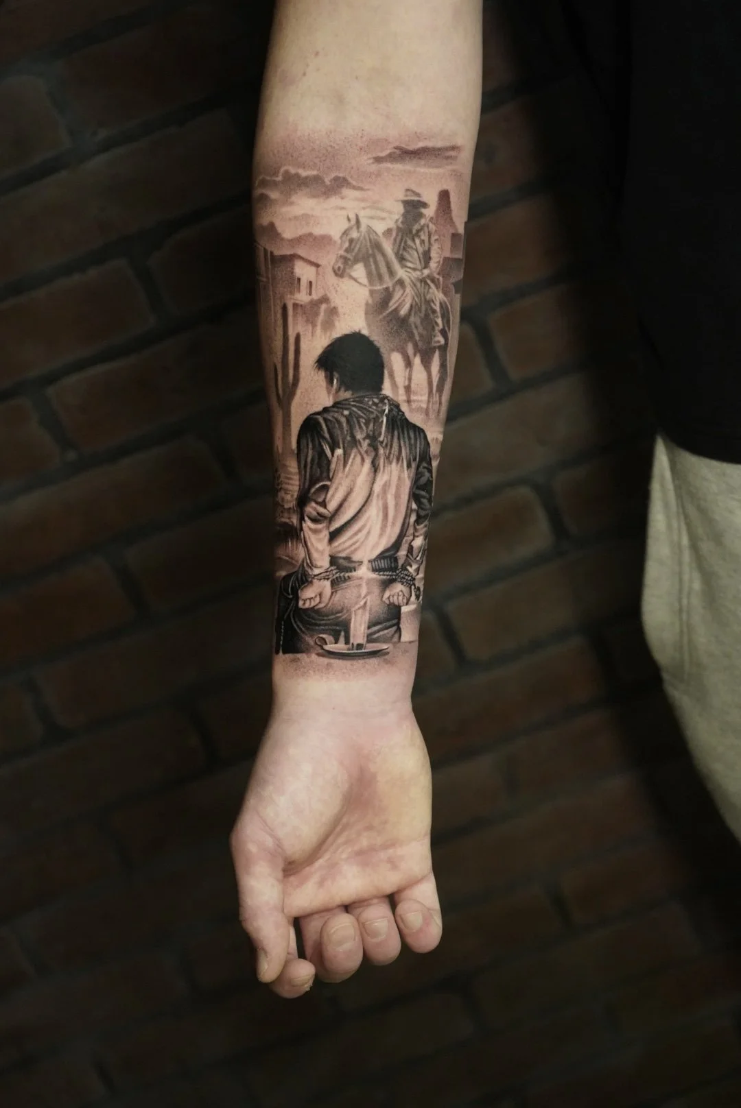 Realistic black and gray tattoo on inner forearm depicting a cowboy on horseback in a western town scene with buildings and a cloudy sky.