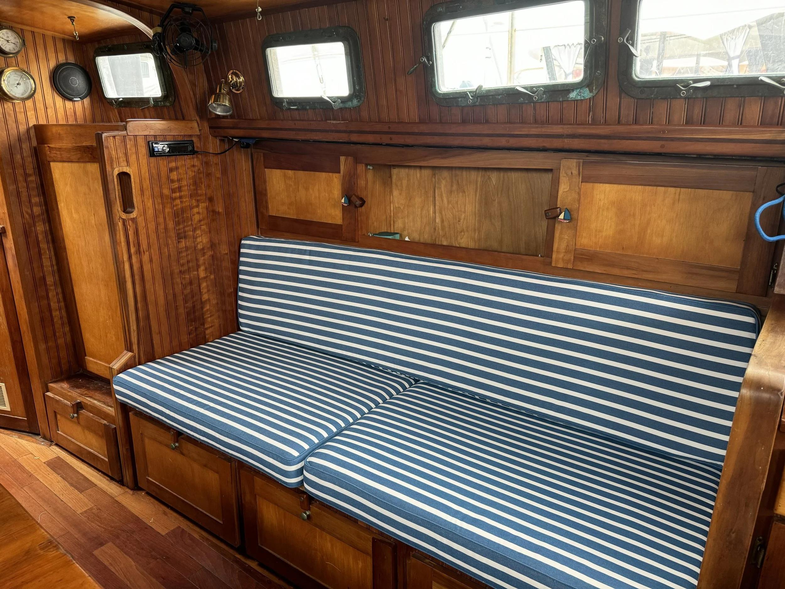 Interior of a wooden boat cabin with a striped blue and white cushion sofa, small drawers underneath, wooden panel walls, and several small windows. Searching for boat covers Sydney, boat covers Port Hacking, or Shellharbour boat covers you can rely