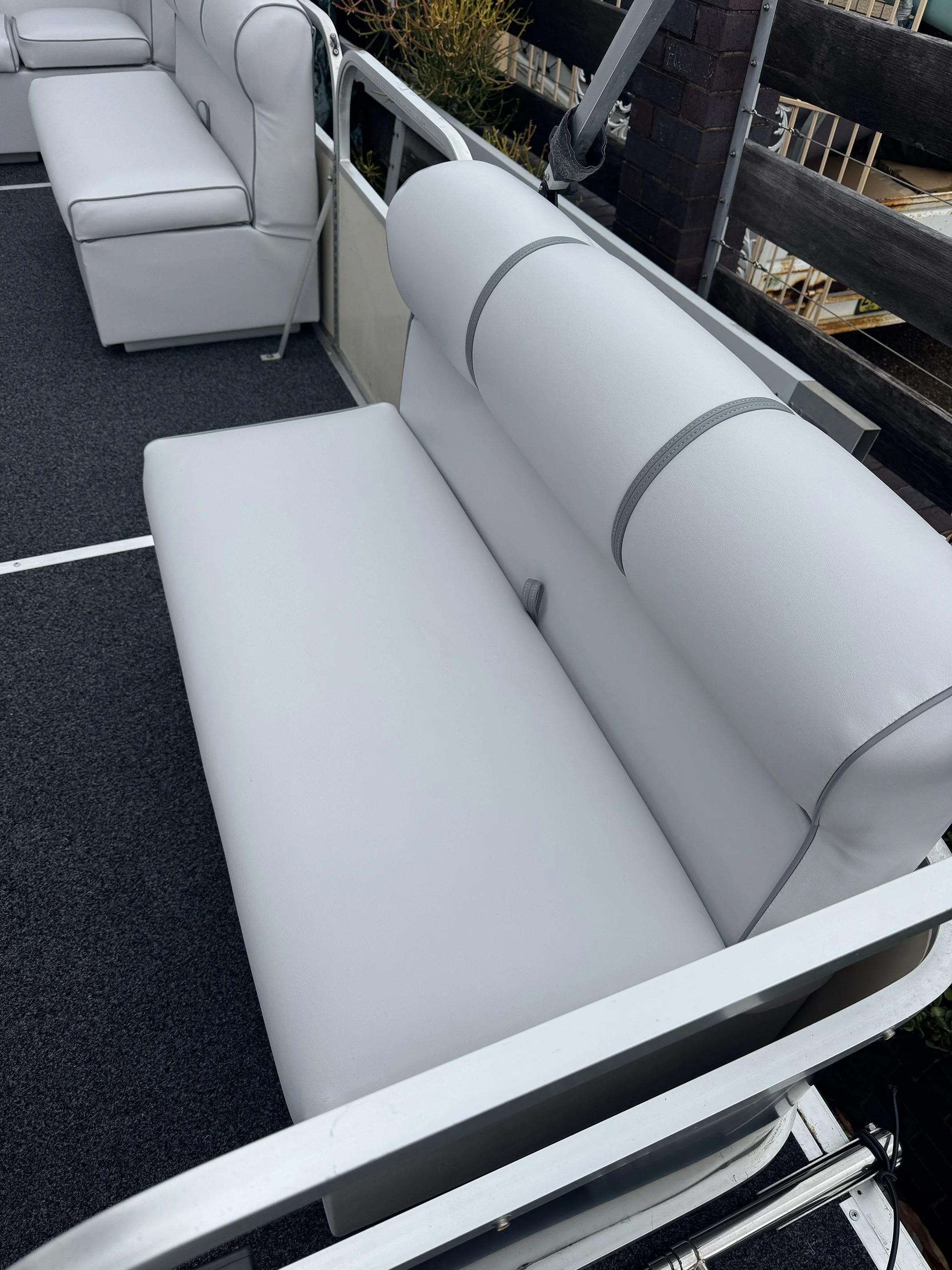 White cushioned seats on a boat deck, with a railing and some plantings in the background. From bimini tops, dodgers, and yacht dodgers to boom bags, boat clears and marine clears, every product is made using durable marine-grade canvas covers built