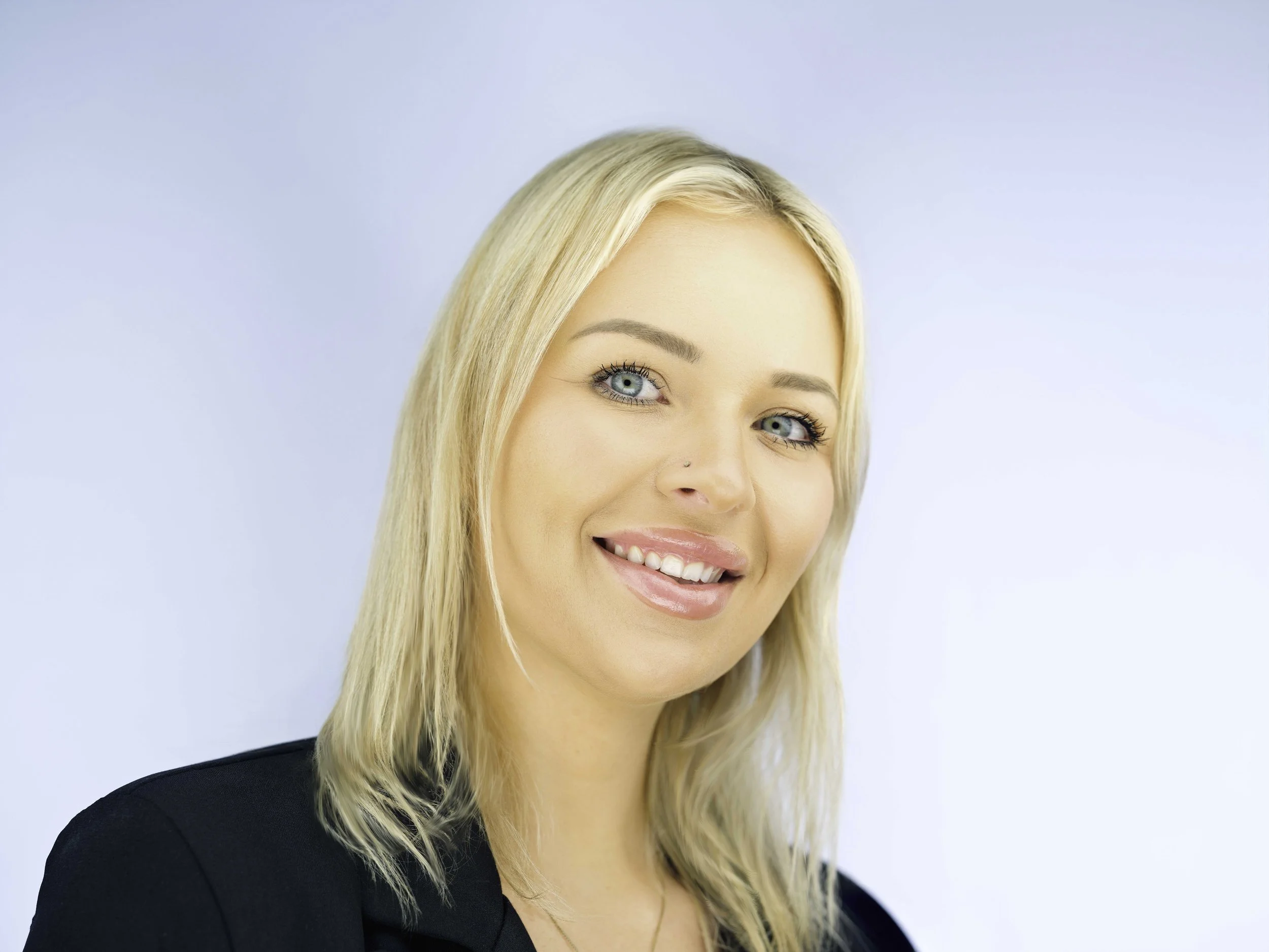 A woman with long blonde hair, blue eyes, wearing a black top, smiling against a light blue background.