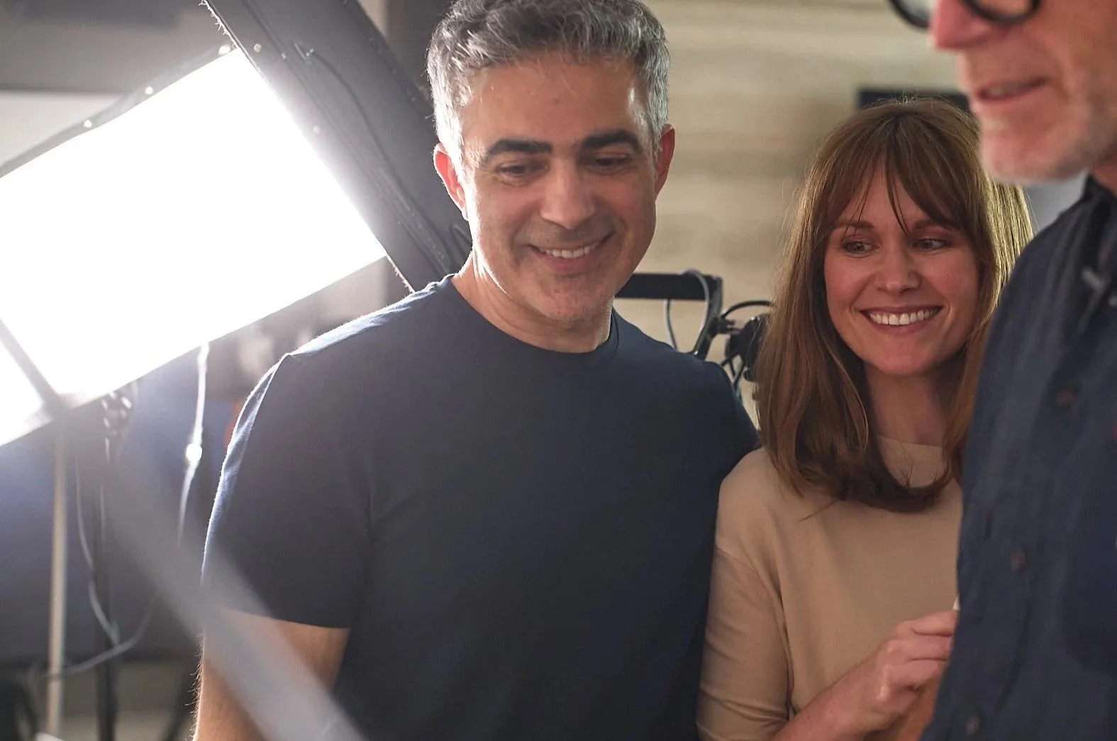 Three people smiling and looking at each other, standing close together in a well-lit indoor setting.