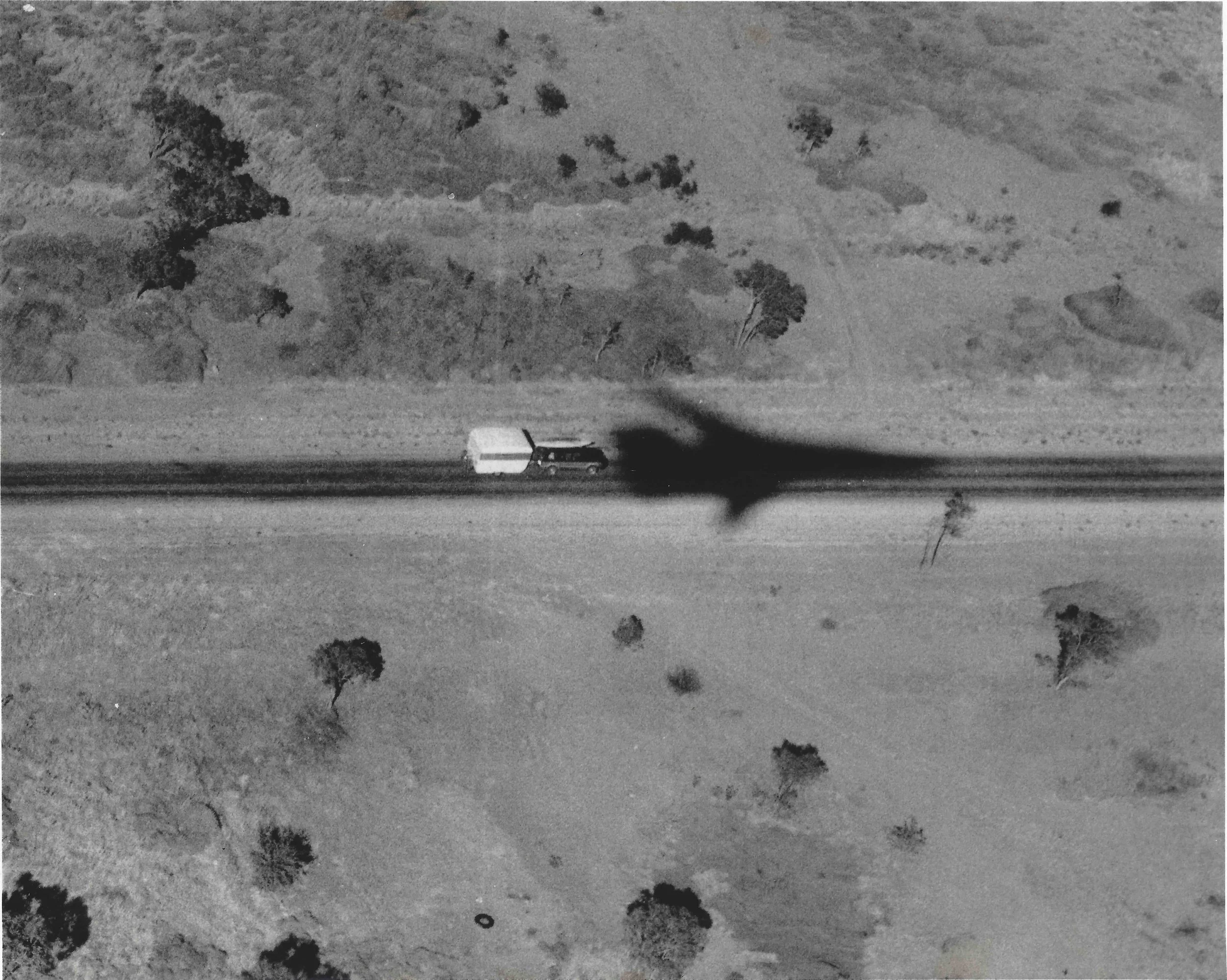 Black and white aerial photo of a rural landscape with trees, fields, and a single vehicle shadowed by an F111 aircraft