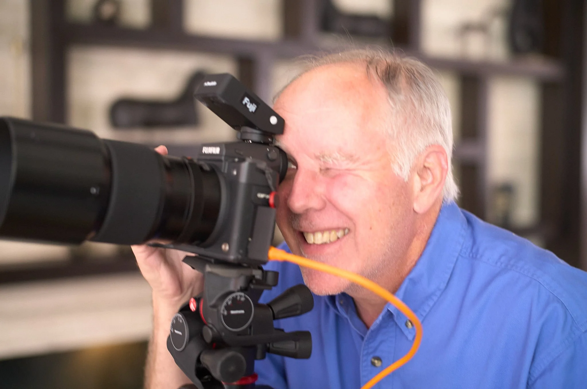 A man with gray hair, smiling, looking through a camera's viewfinder, wearing a blue shirt, with a blurred background.