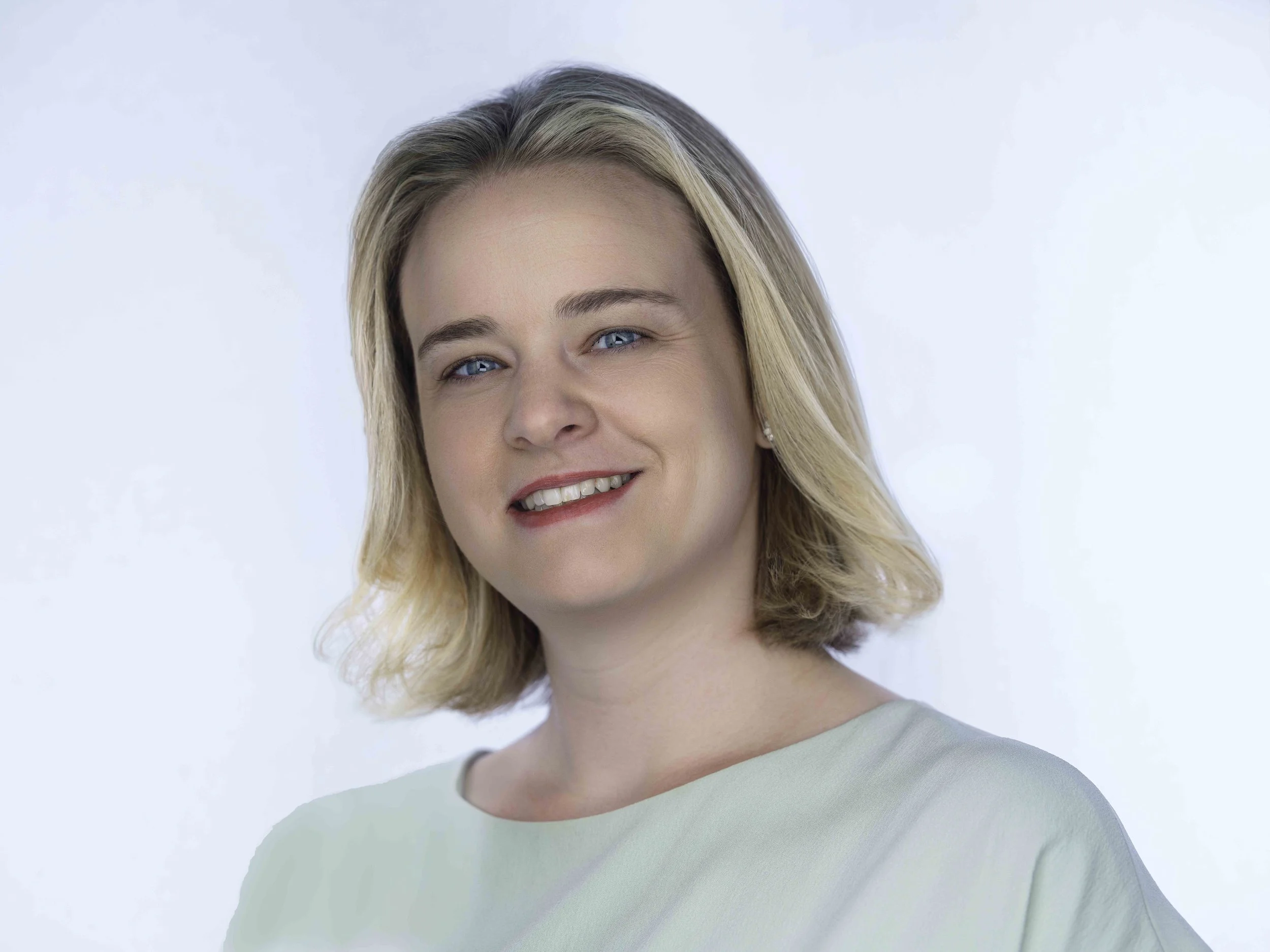 A smiling woman with blonde hair, blue eyes, and light skin wearing a mint green top, standing against a light blue background.