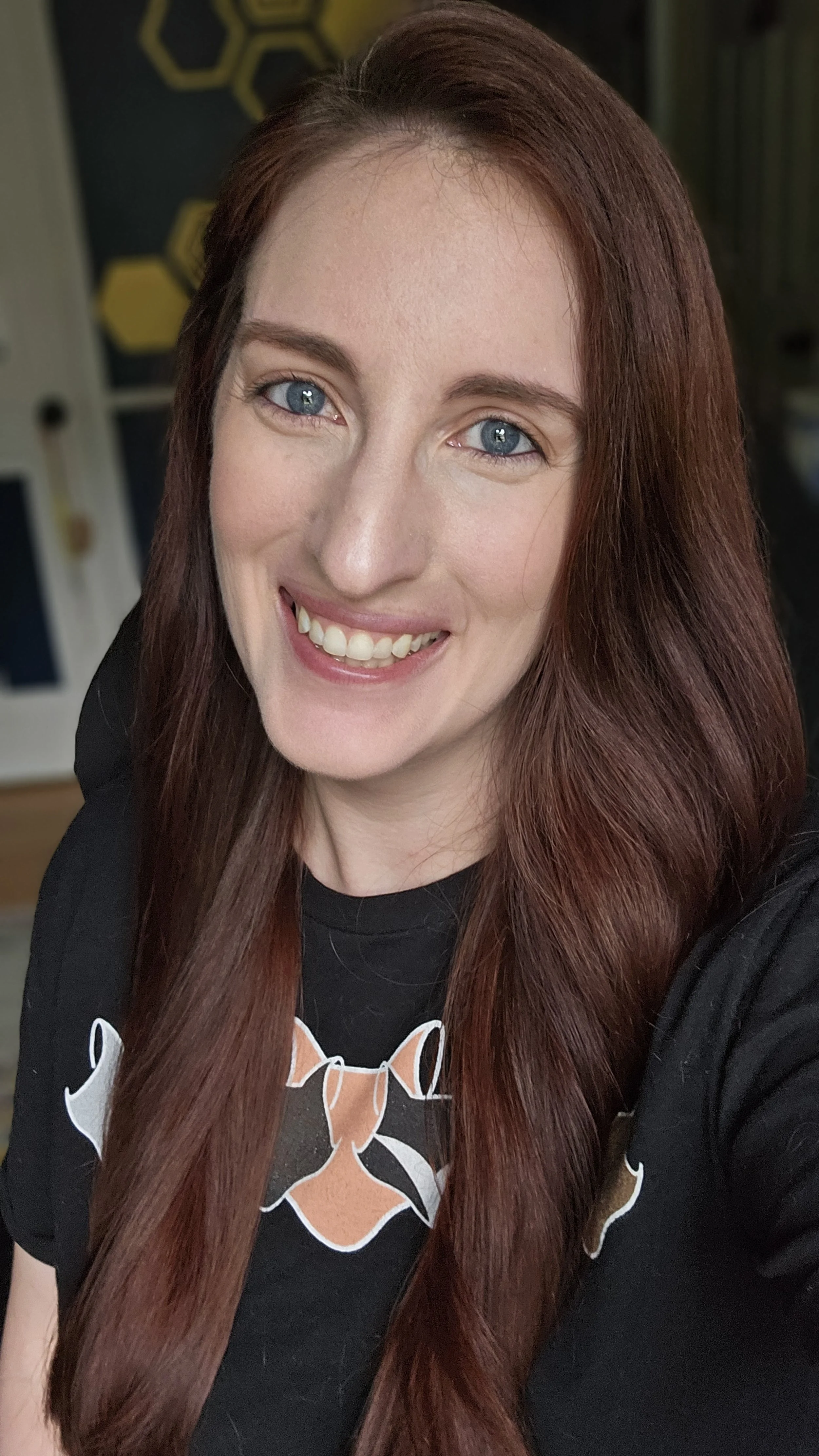 A woman with long red hair and blue eyes smiling at the camera in an indoor setting with a dark wall background.
