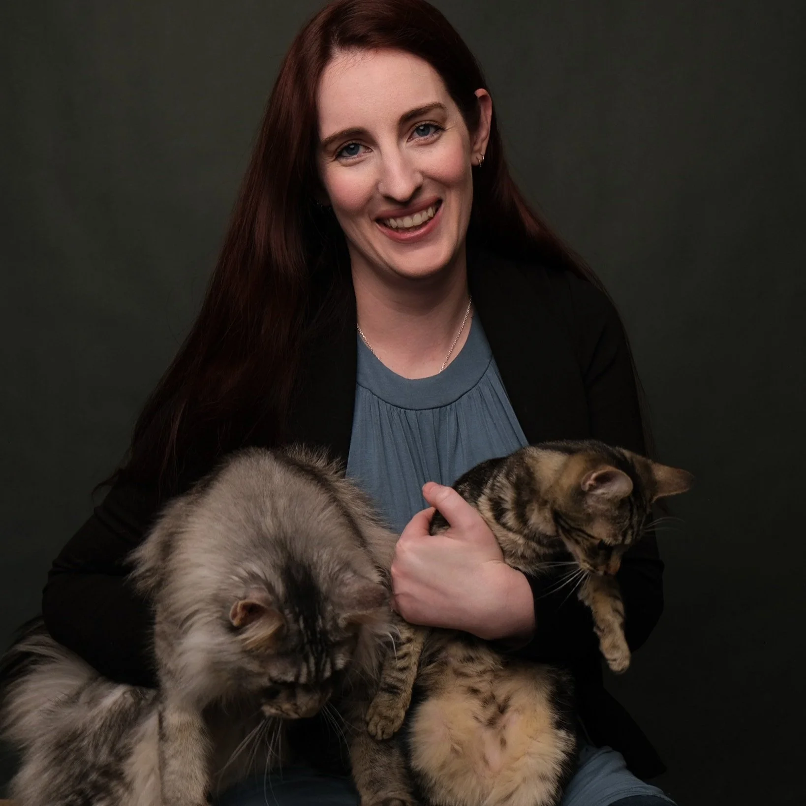 A smiling woman with long red hair holding two cats, one in each arm, in front of a dark background.