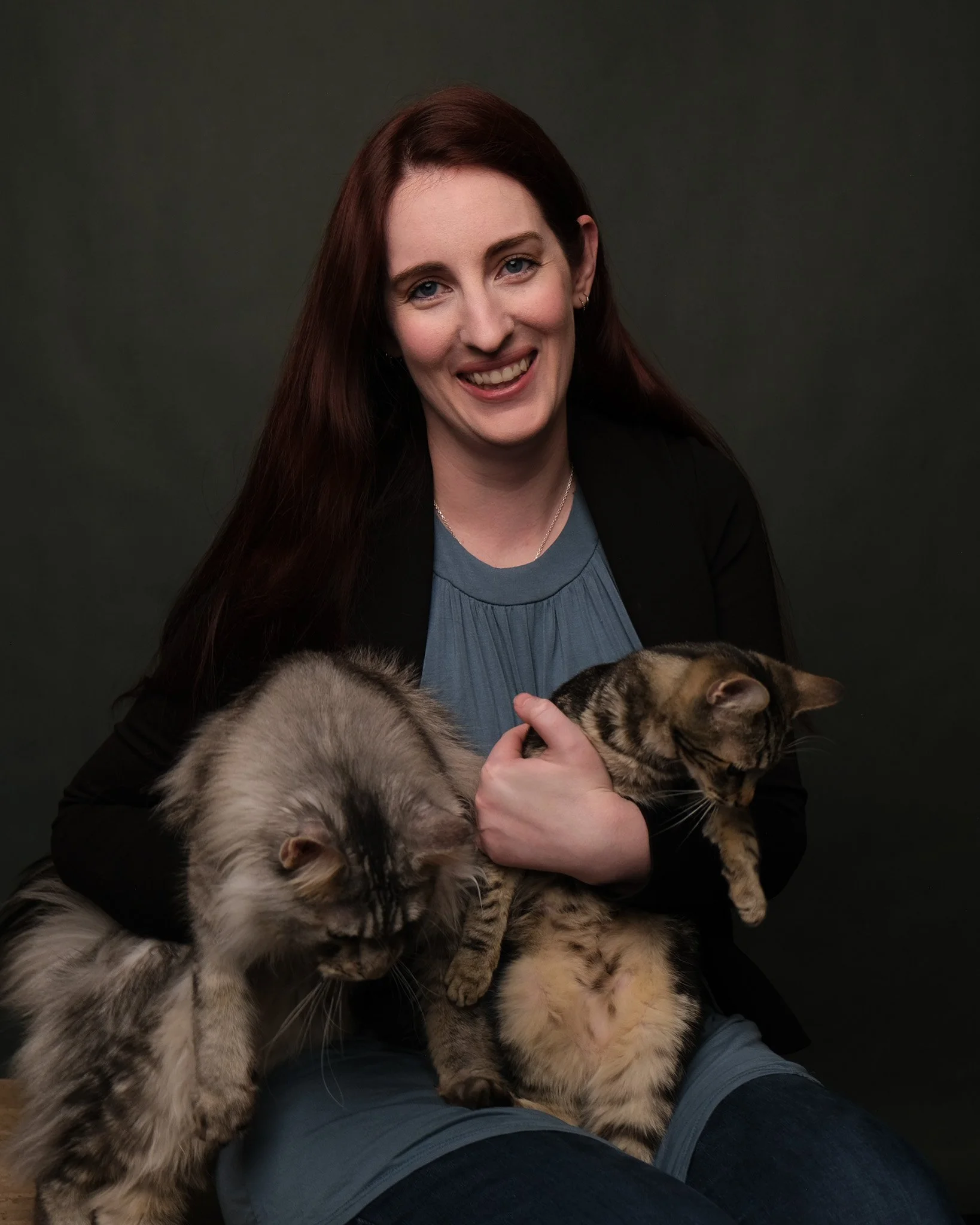 A woman with long red hair, smiling, holding two cats, one on her lap and one in her arm, against a dark background.