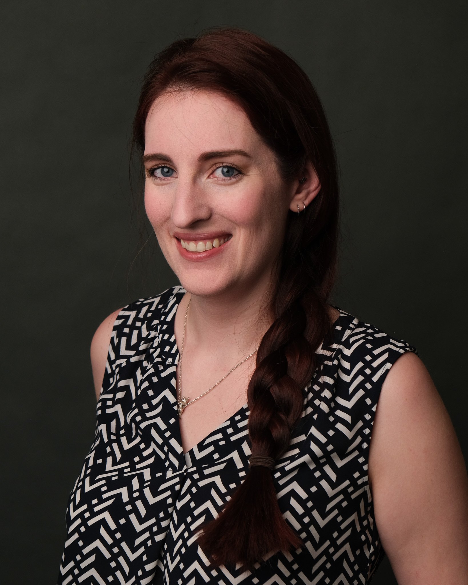 A woman with long, dark red braided hair smiling, wearing a sleeveless blue and white geometric patterned top, against a dark grey background.