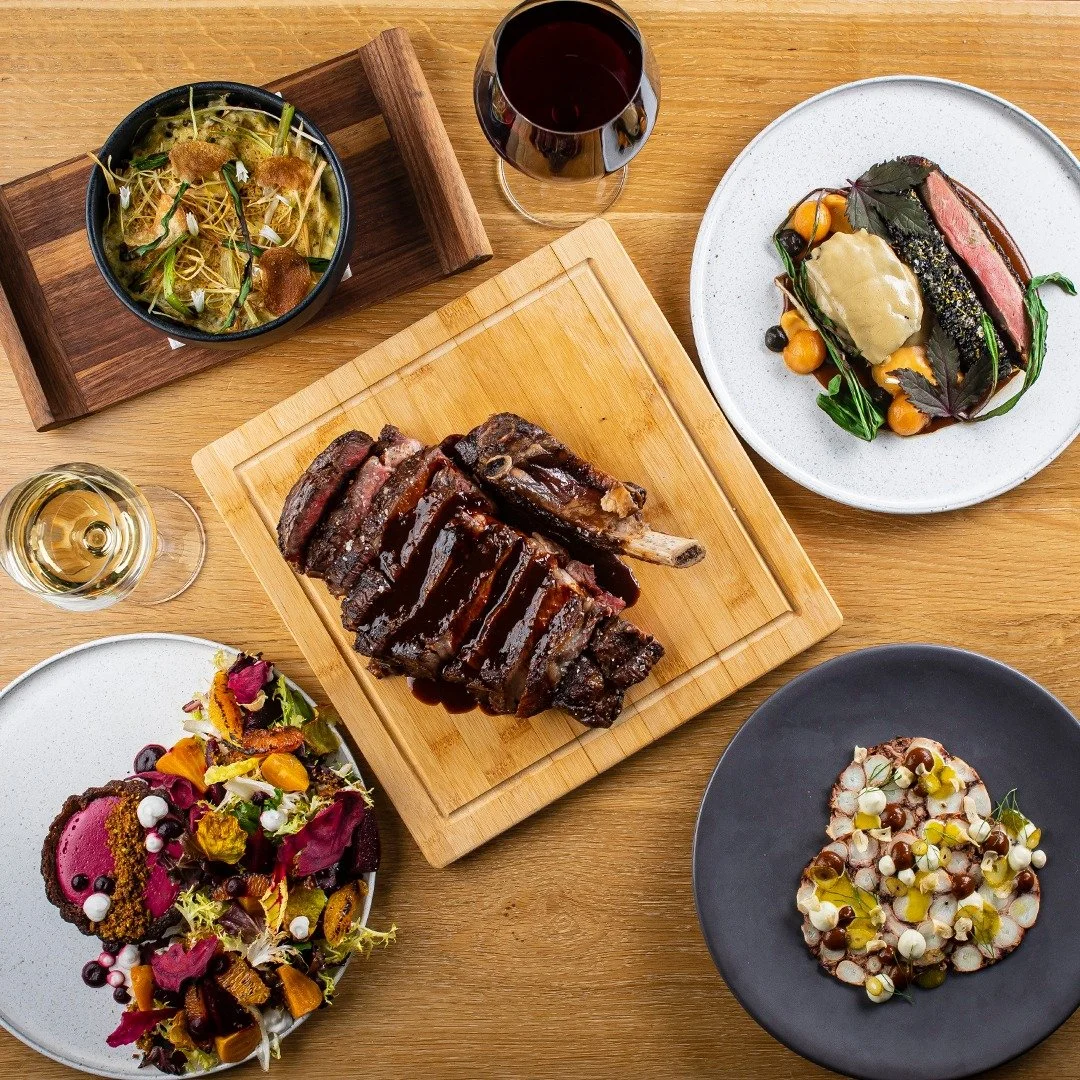 A gourmet dinner with a rack of ribs, two glasses of red and white wine, and several artfully plated dishes on a wooden table.