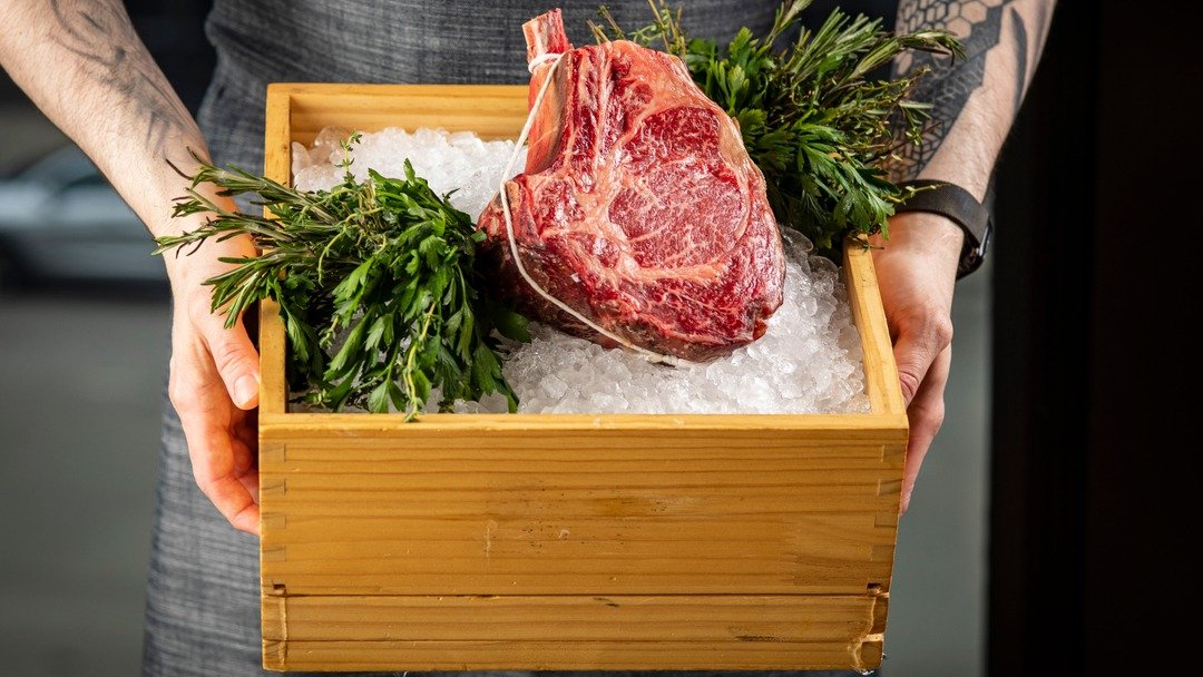 Justin holding a wooden box with ice, fresh herbs, and a large cut of raw beef steak.
