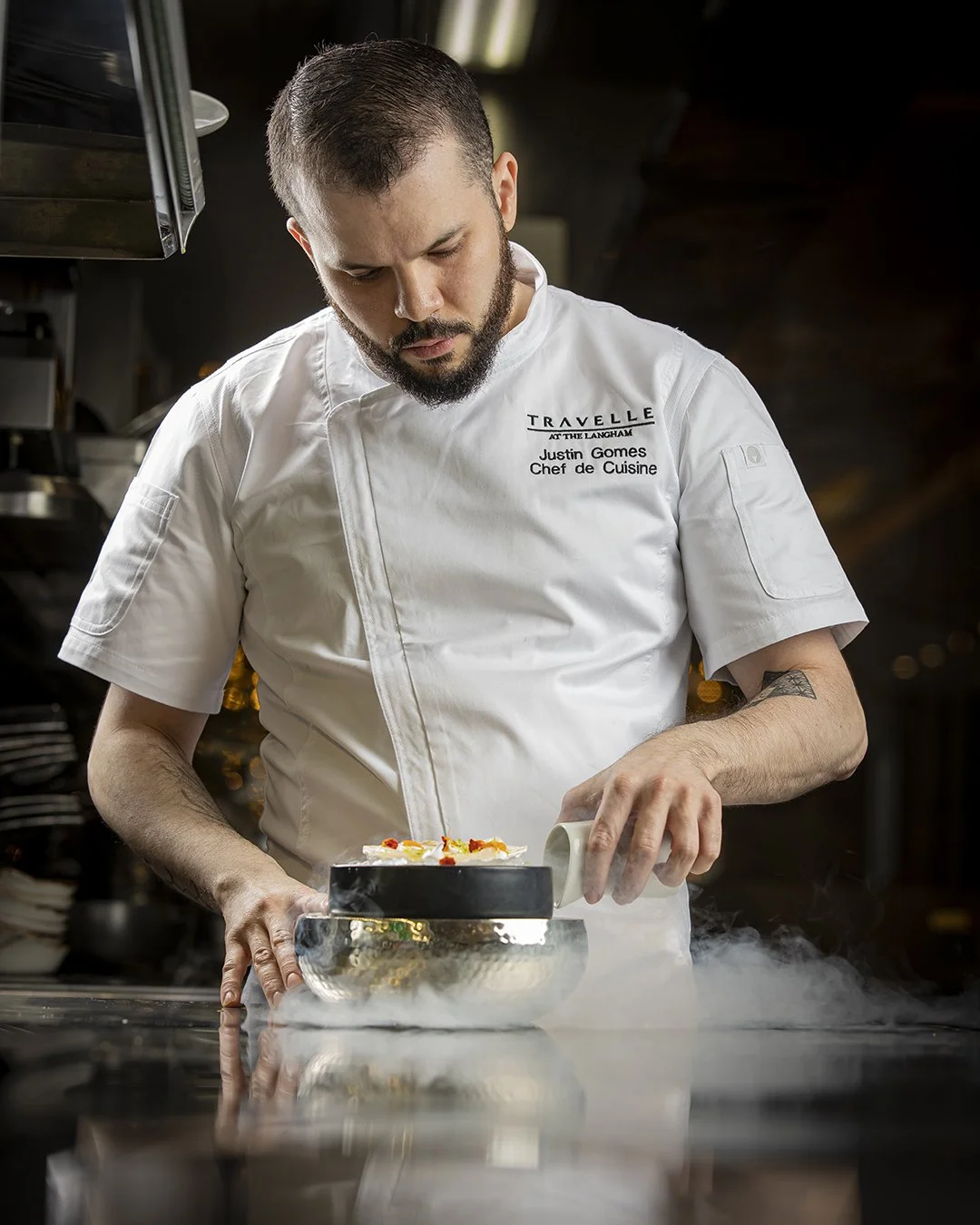 A male chef in a white uniform with the name tag 'Justin Gomes, Chef de Cuisine' preparing food in a professional kitchen, with steam rising from the dish.