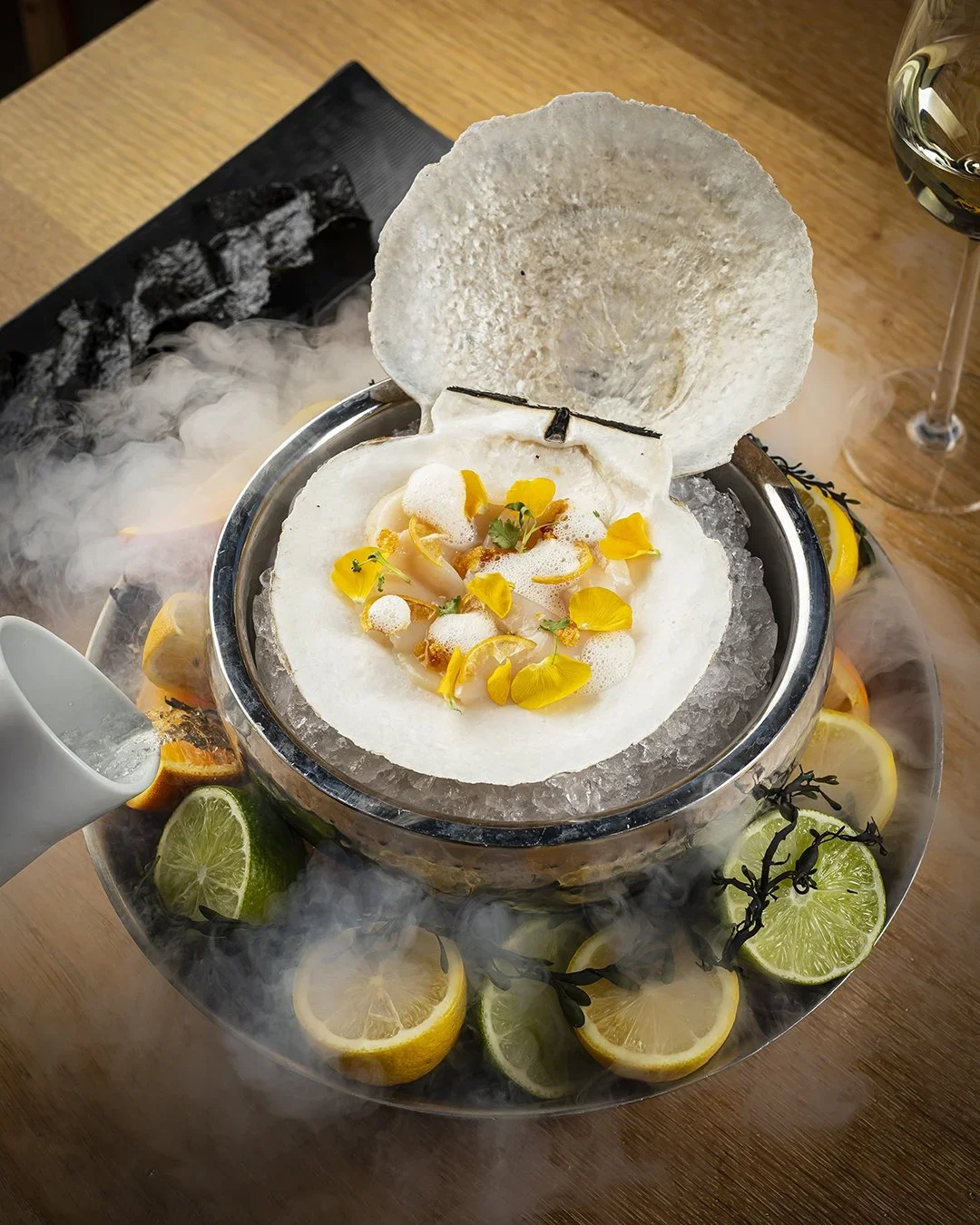 A seafood dish served in a shell, garnished with yellow flower petals and herbs, surrounded by lemon and lime slices, with dry ice and smoke effects.