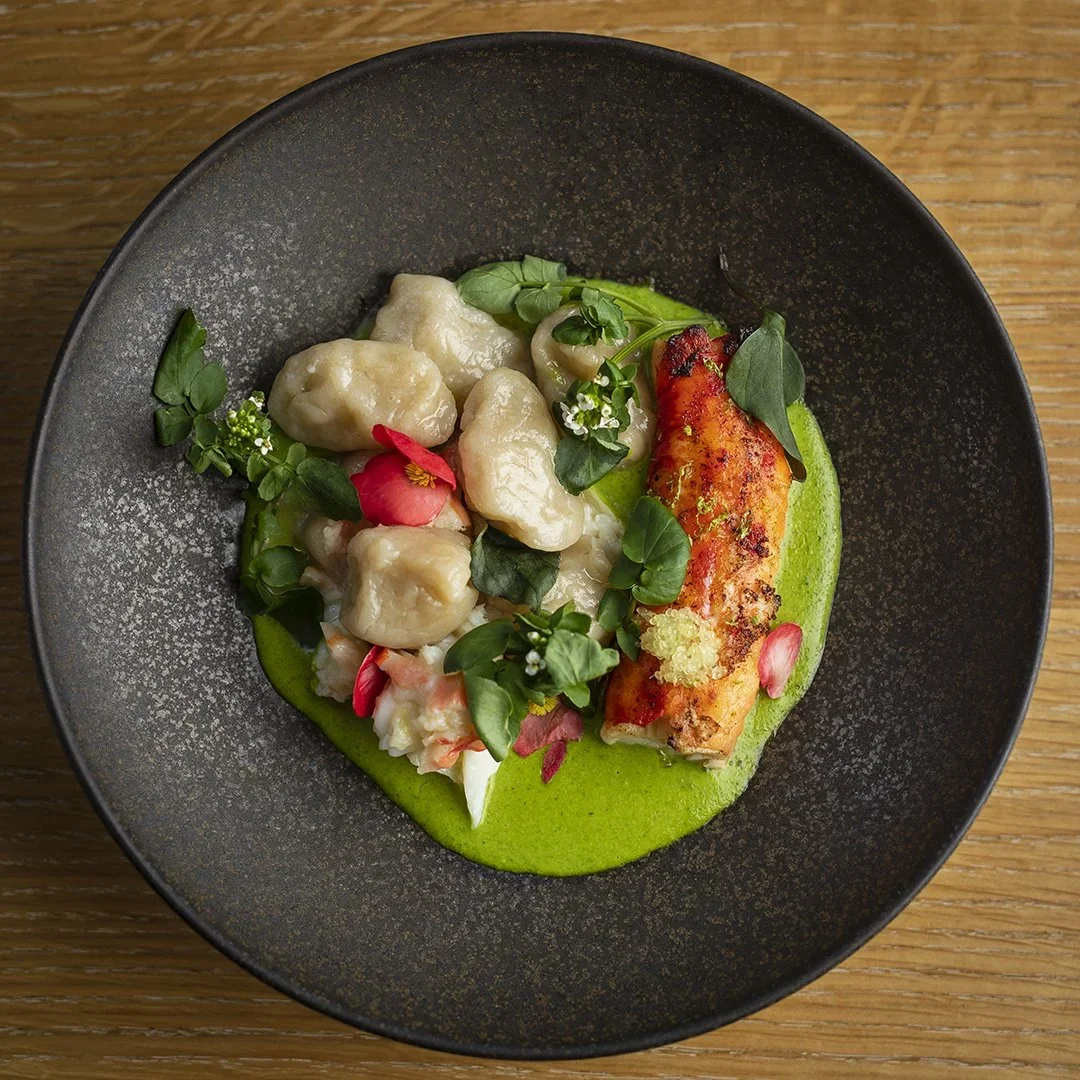 A black plate with gnocchi, king crab pieces, green curry sauce, garnished with herbs, edible flowers, and petals.