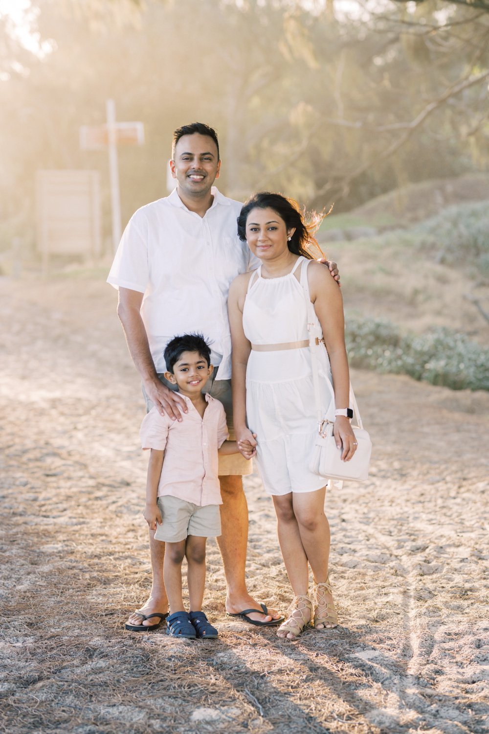 A family of four standing outdoors on a dirt path with trees in the background, holding hands. It is sunny with warm lighting.