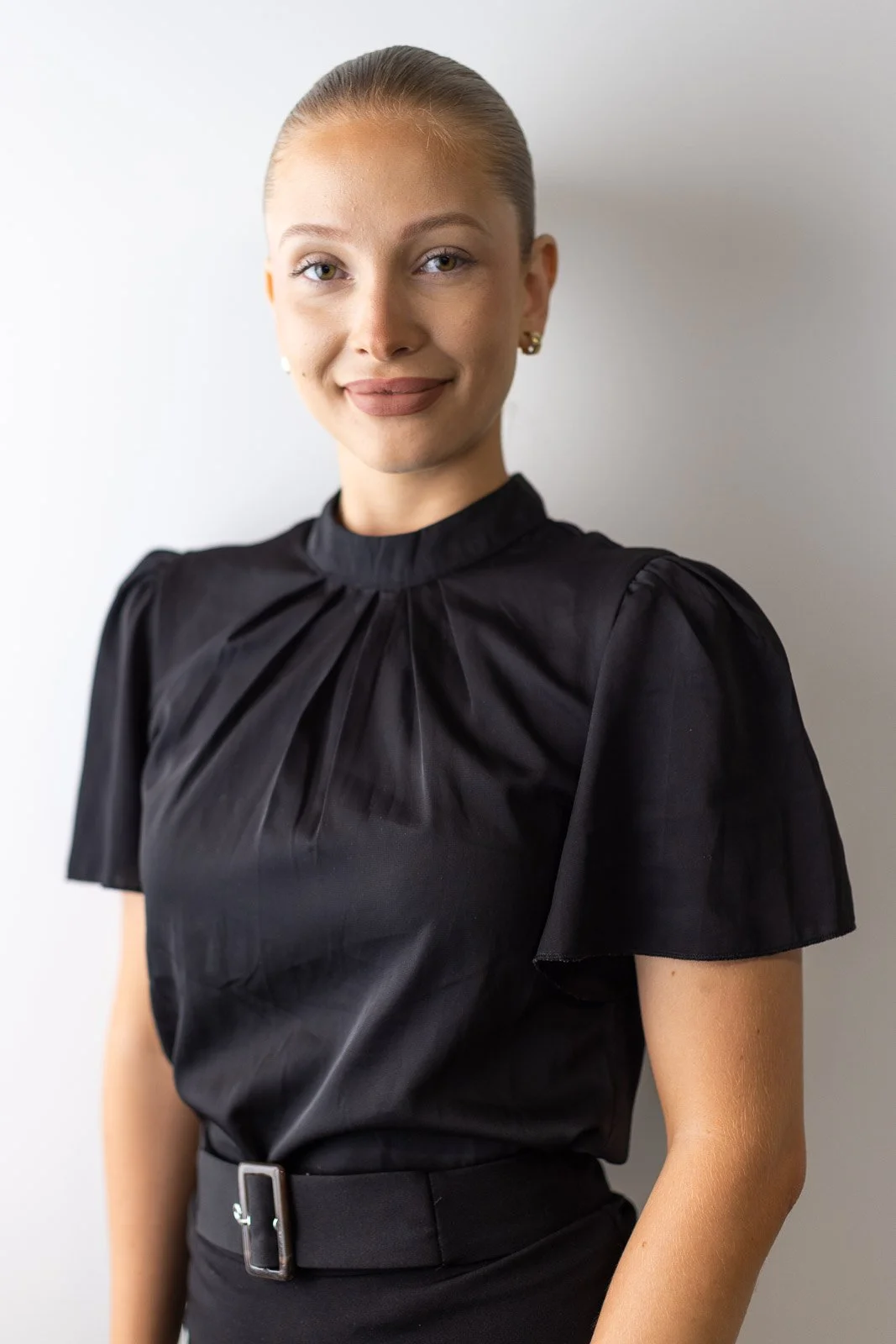 A woman with blonde hair tied back, wearing a black blouse with pleats and puffed sleeves, and earrings, standing against a plain light gray wall.