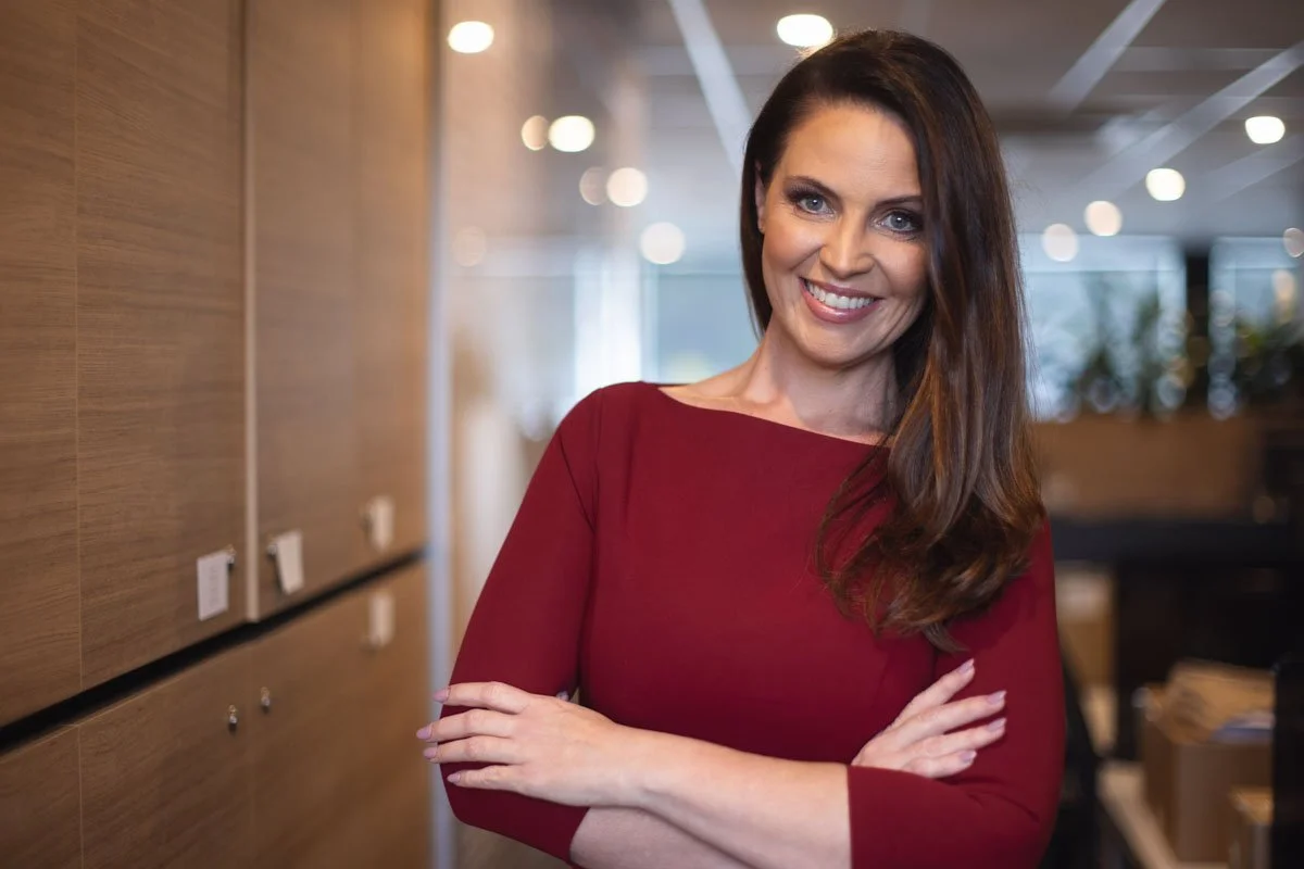 A woman with long brown hair wearing a red dress, smiling with crossed arms in an office setting.