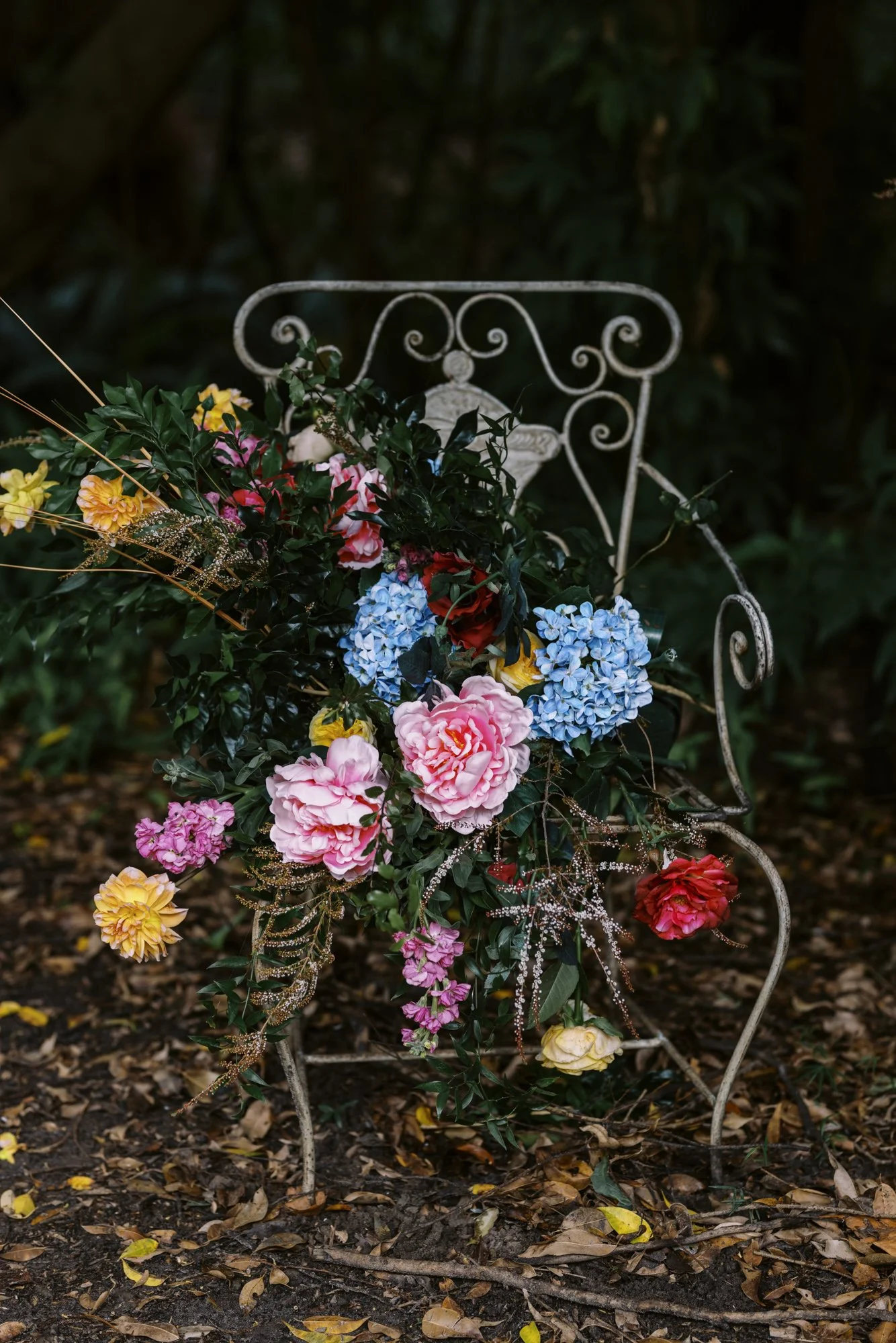 Iron garden bench with a large bouquet of colorful flowers including pink peonies, blue hydrangeas, yellow, red, and purple blooms, and green foliage, set outdoors on leaf-covered ground.