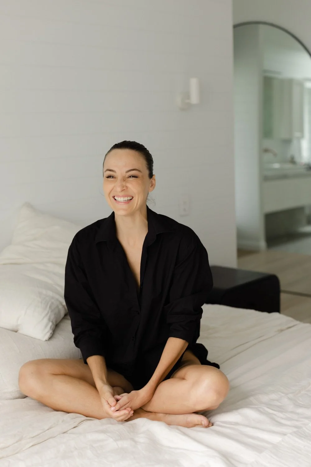 A woman sitting cross-legged on a bed, smiling and laughing, wearing a black shirt in a modern, minimalistic bedroom.