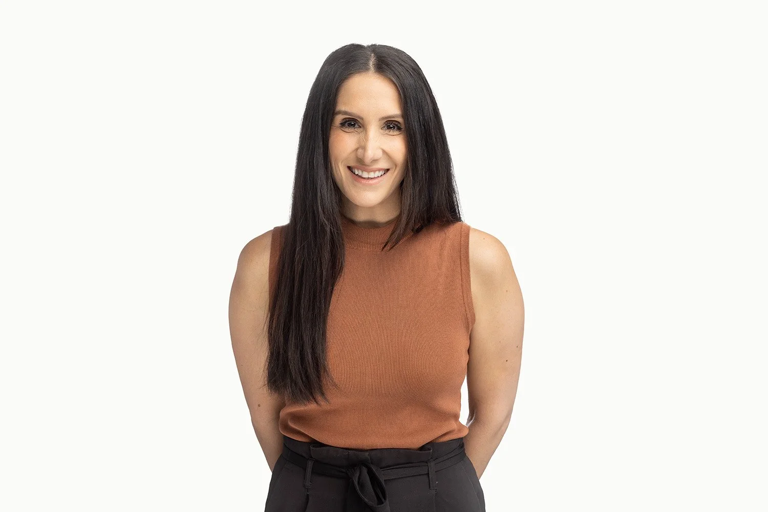 A woman with long dark hair smiling, wearing a sleeveless brown top and black pants against a white background.