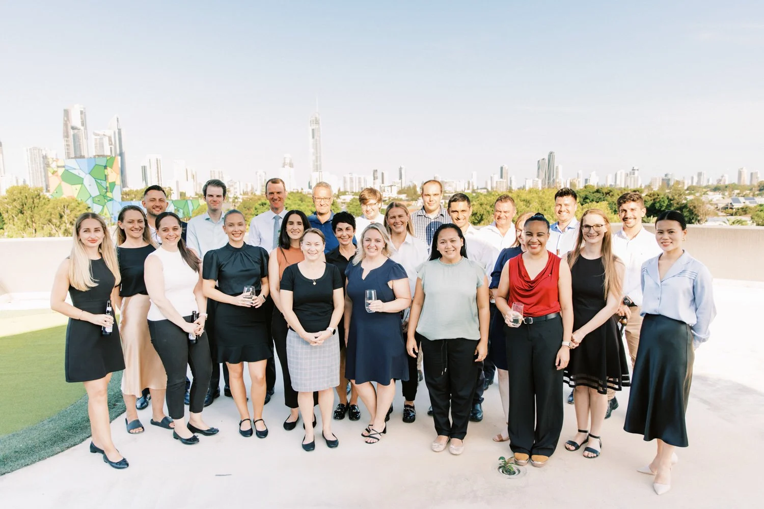 Group of 20 diverse professionals gathered outdoors on a patio with a city skyline in the background, some holding drinks, smiling for a group photo.