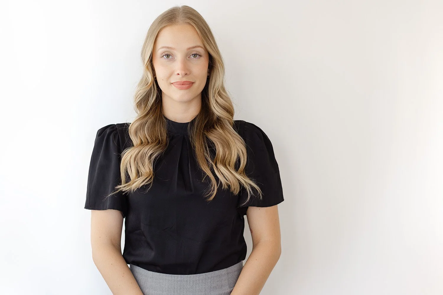 Young woman with long wavy blonde hair wearing a black top and gray skirt standing against a white wall.
