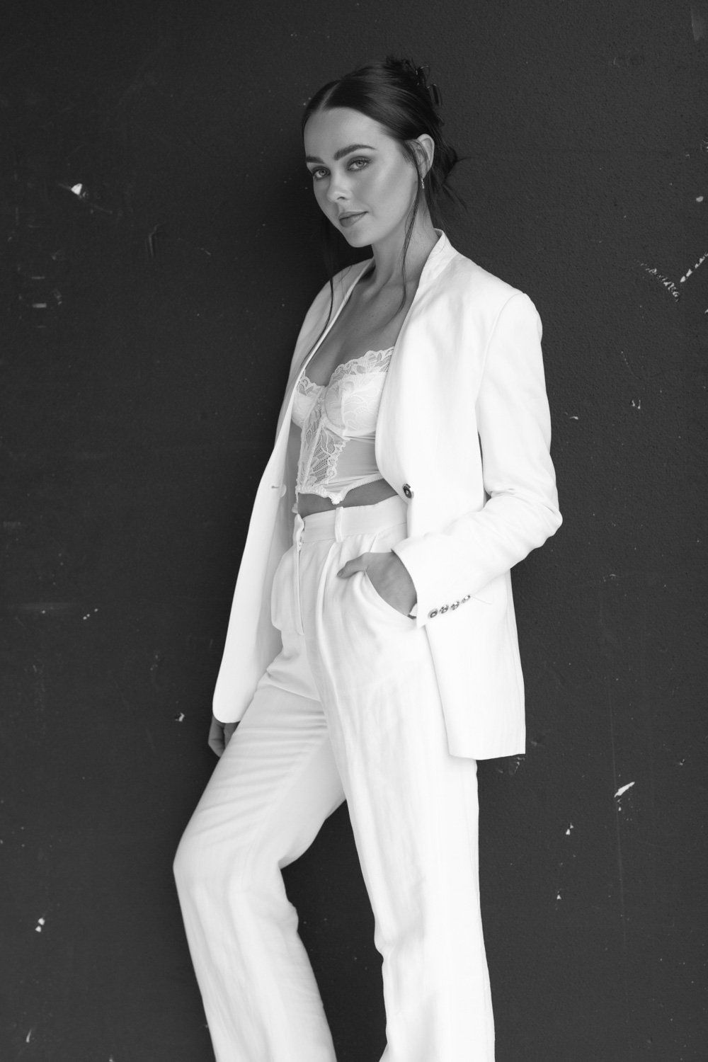 A woman in a white pantsuit and lace top stands against a dark wall, posing with one hand in her pocket, looking at the camera.