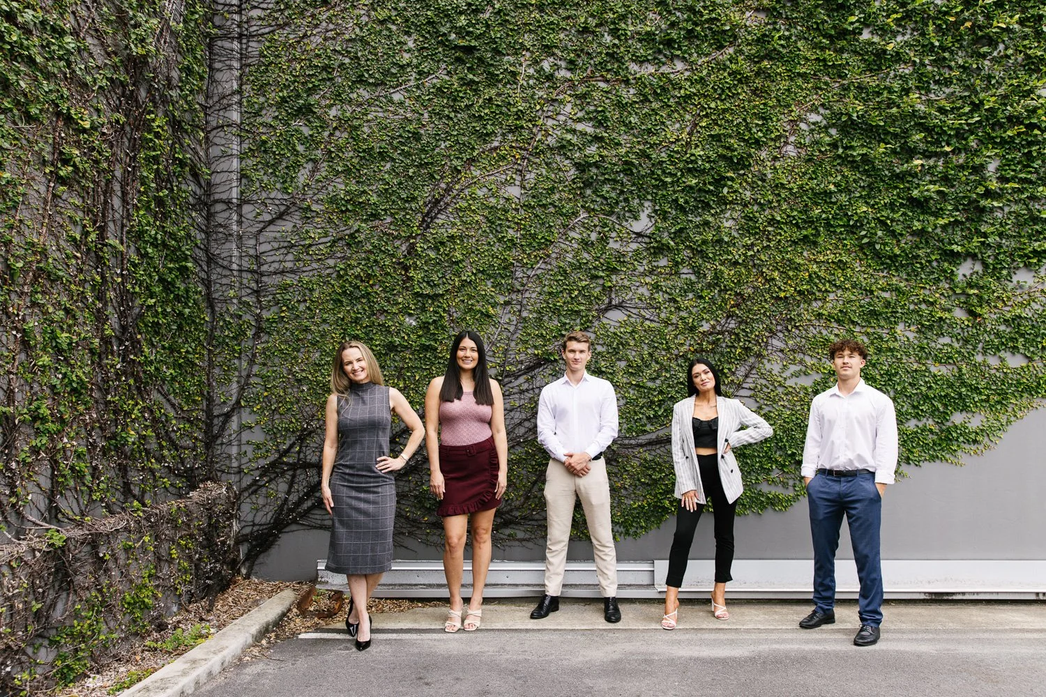 Group of five young professionals standing outdoors in front of a gray wall covered with green ivy, dressed in business casual attire.