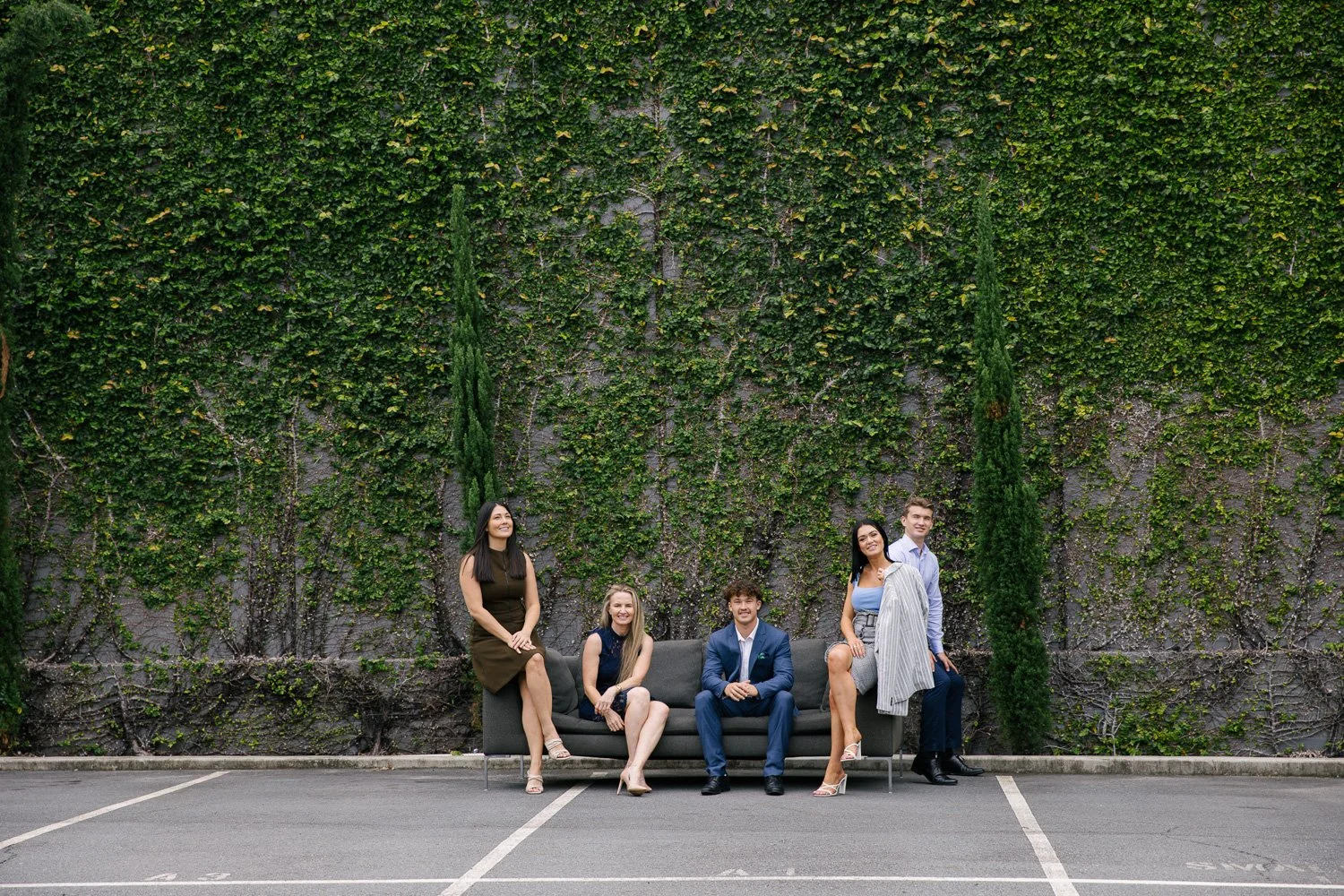 Six people sitting and standing in front of a green ivy-covered wall in a parking lot, including a sofa and all dressed in business casual and formal attire.