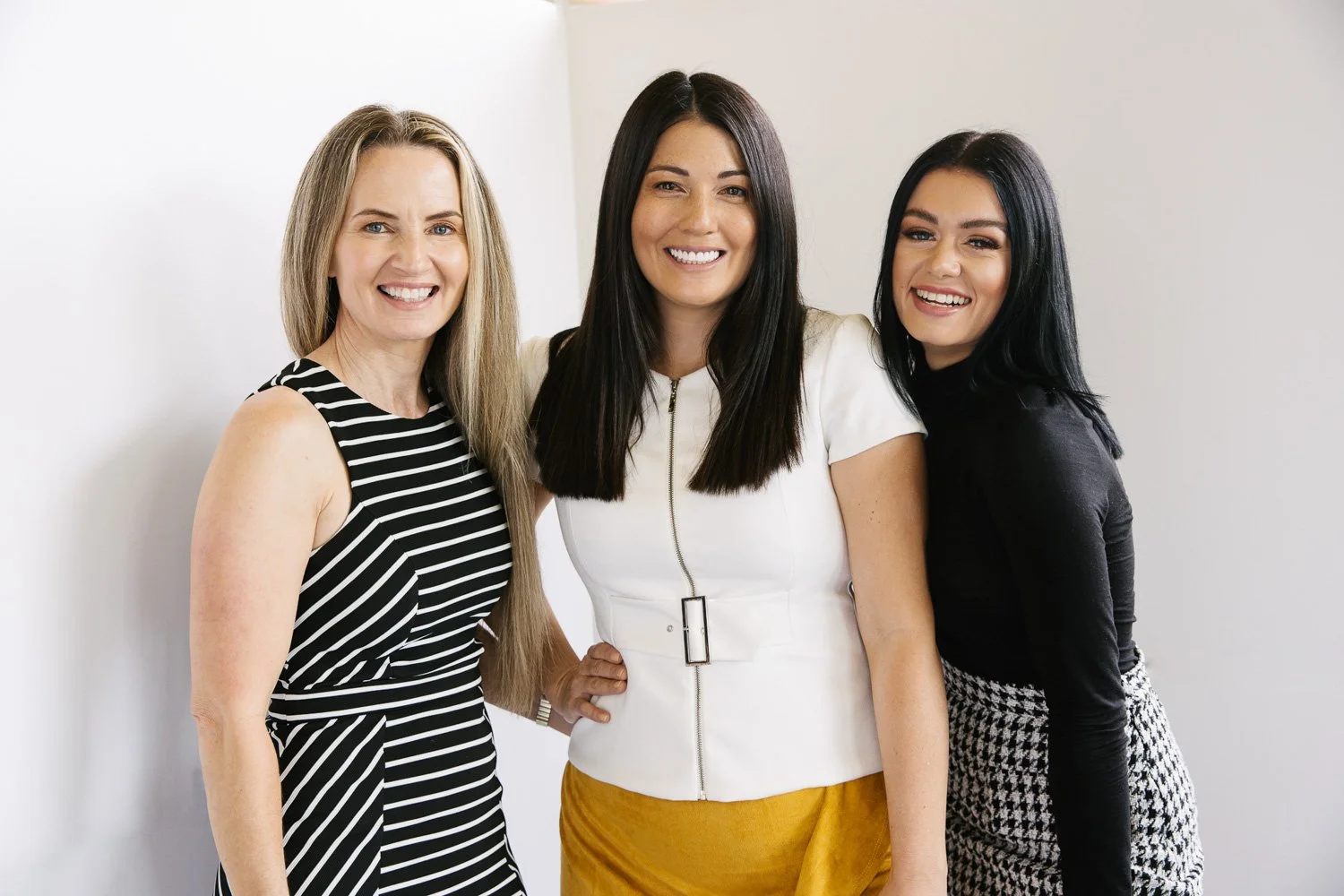 Three women smiling and standing together against a plain white background.