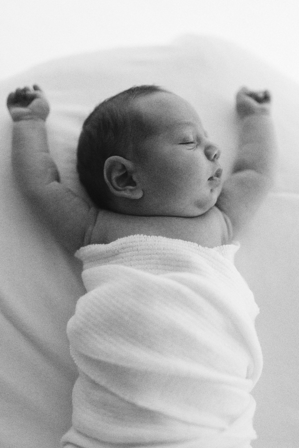 Black and white photo of a newborn baby sleeping with arms stretched upwards, swaddled in a white blanket.