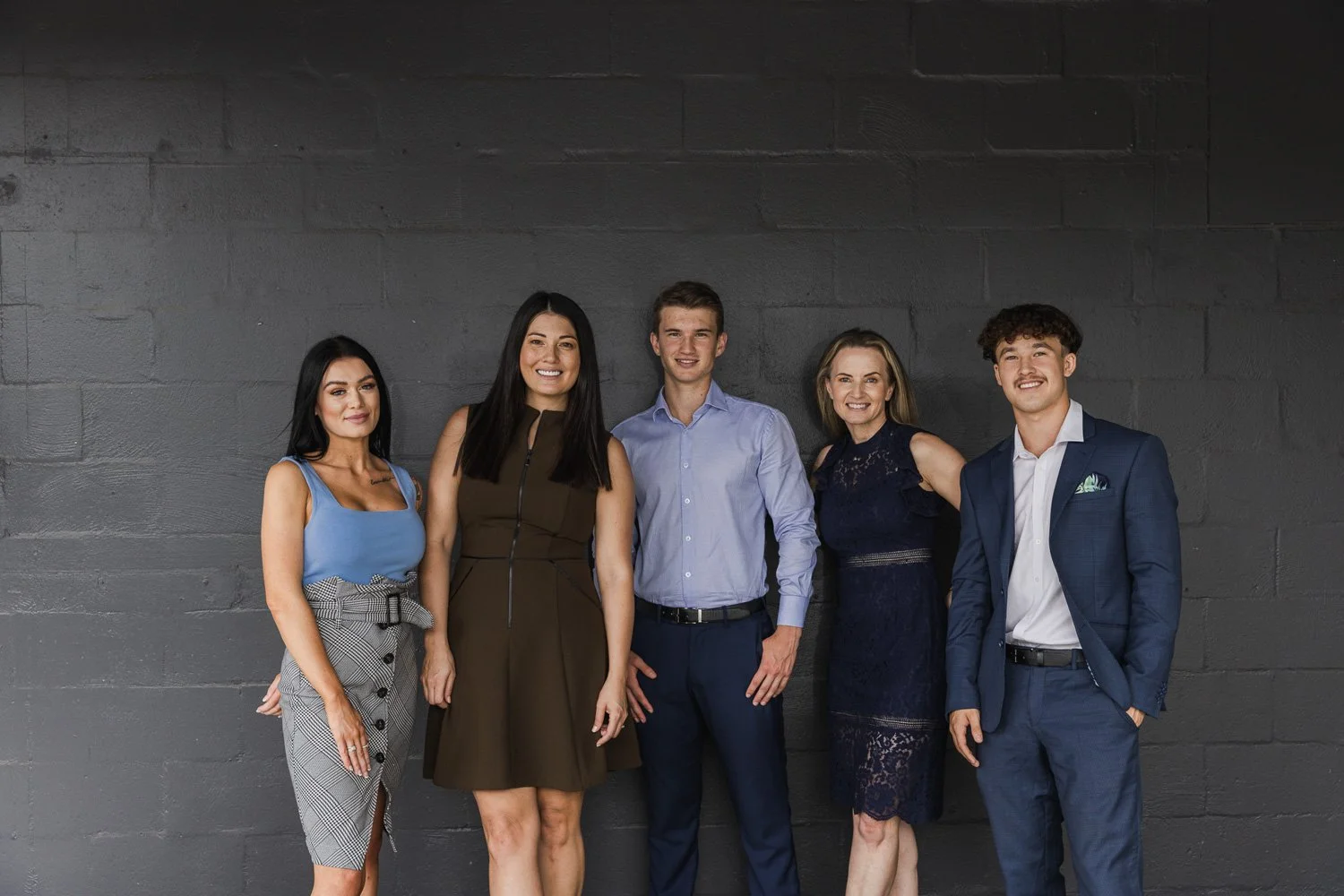 Group of five professionally dressed people standing against a dark gray brick wall, smiling at the camera.