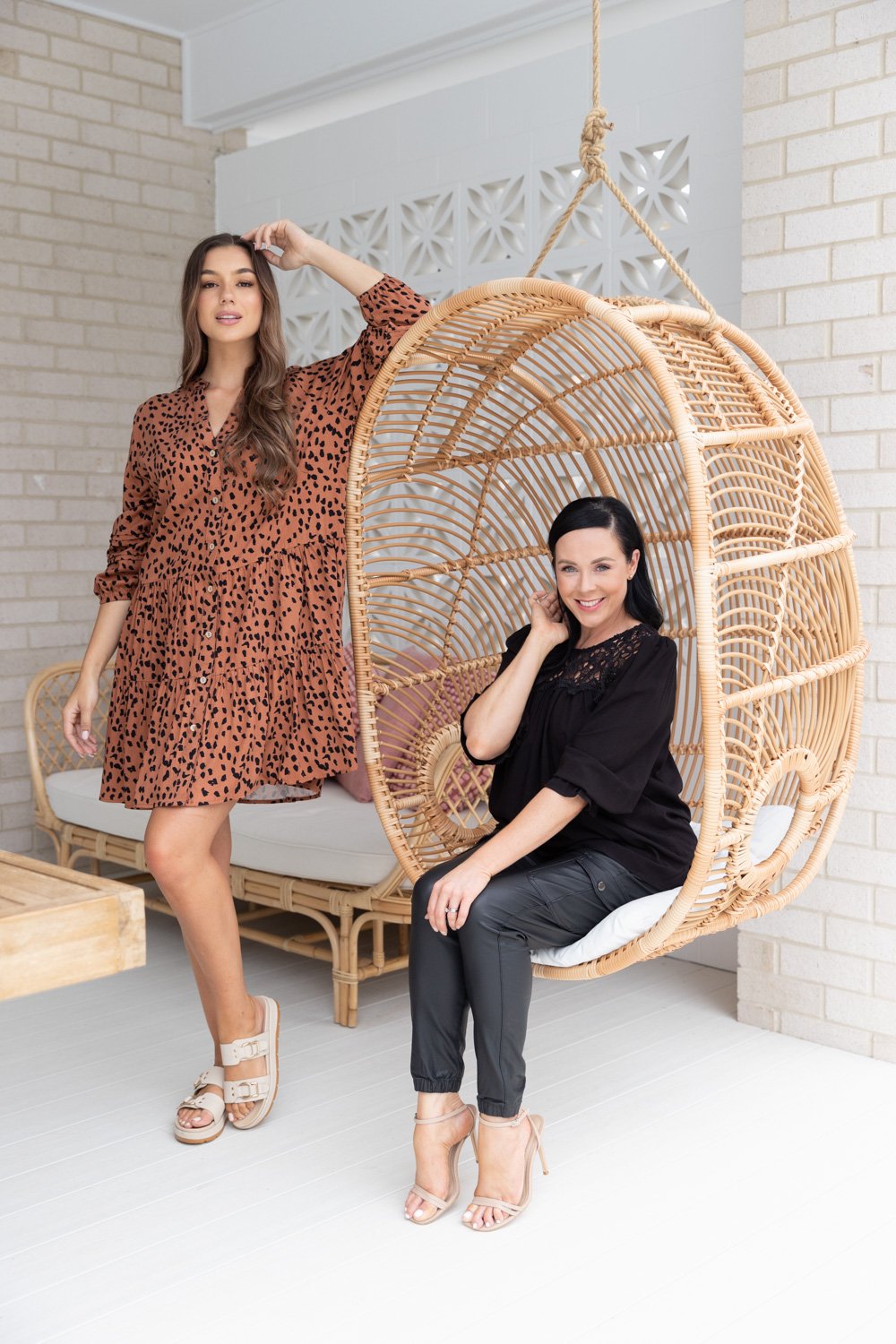 Two women posing inside and next to a hanging wicker chair in a room with white brick walls. One woman is standing wearing a leopard print dress, and the other is sitting in the wicker chair wearing a black top and leather pants.