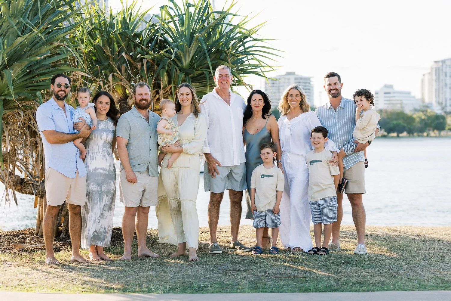A large family group on a beach near water with buildings in the background, dressed in light, summery clothing, smiling and posing together.