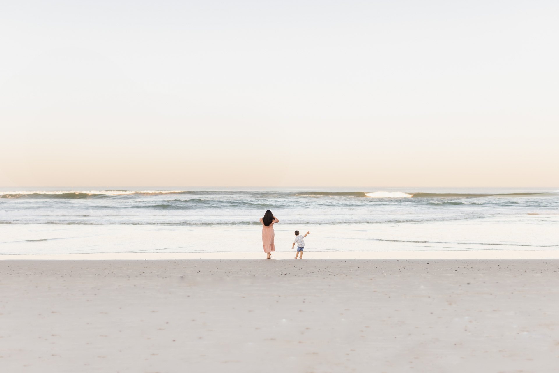 A woman and a young boy stand on a sandy beach facing the ocean, with the woman holding her hand to her hair and the boy pointing towards the water. The sky is clear and the waves are gently rolling in.