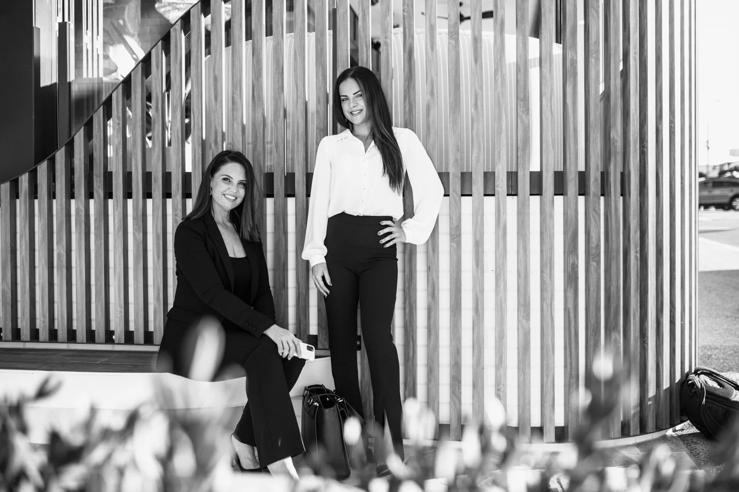 Two women in professional attire posing outdoors in front of a wooden slat wall, one sitting on a bench and the other standing with hand on hip.