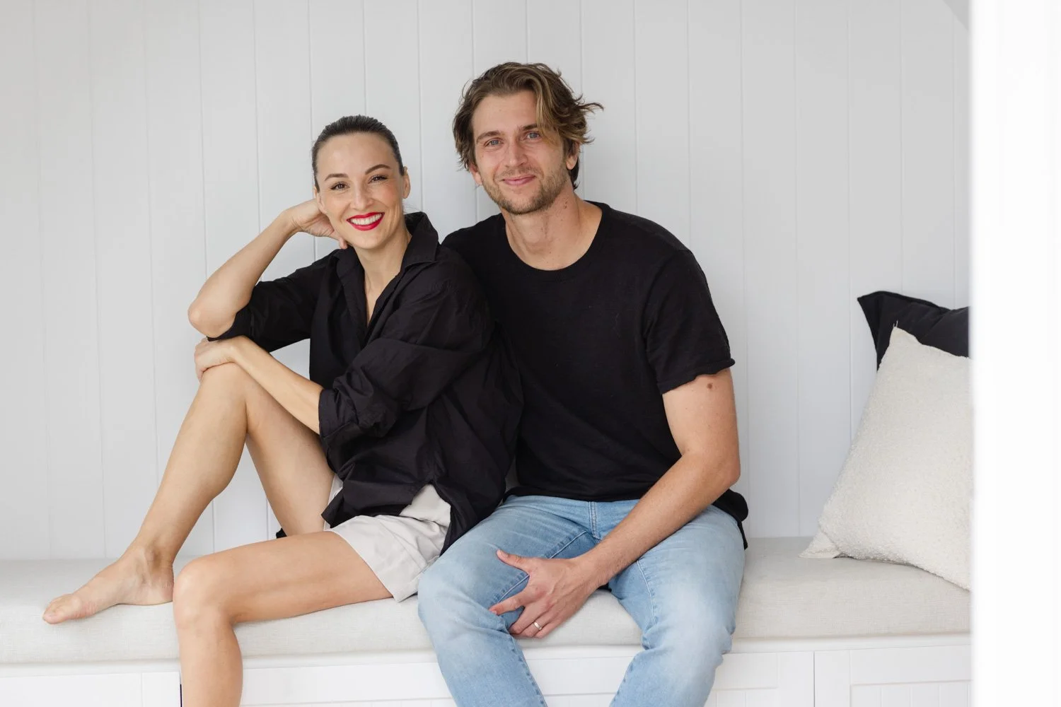 A woman and man sitting on a white bench against a white wall, smiling at the camera. The woman has black hair tied back, wears red lipstick, a black shirt, and beige shorts. The man has wavy brown hair, a beard, a black T-shirt, and light blue jeans