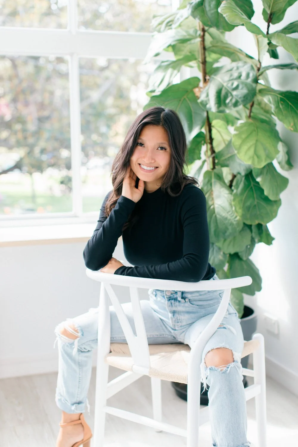 A woman sitting on a white chair near a large window with greenery outside, wearing a black long sleeve top and ripped jeans, smiling at the camera with one hand touching her face.
