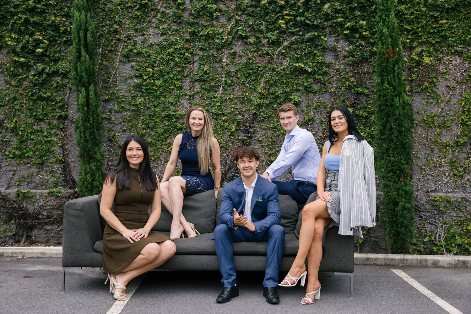 Group of five young adults sitting and standing on a dark gray sofa in front of a green ivy-covered wall outdoors.