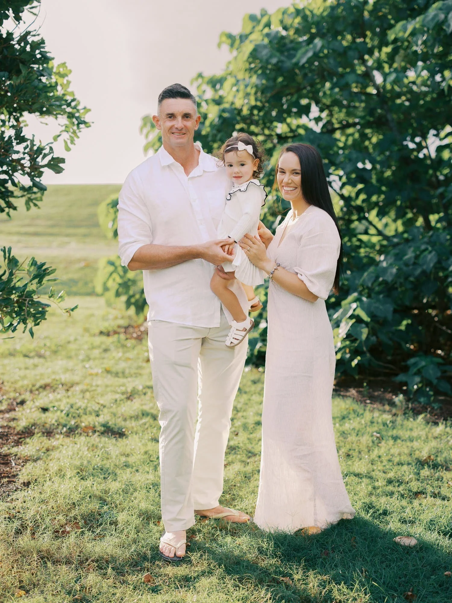 A happy family of three standing outdoors in a lush green park with trees, during daytime. The man, woman, and young girl are smiling and dressed in light-colored clothing.