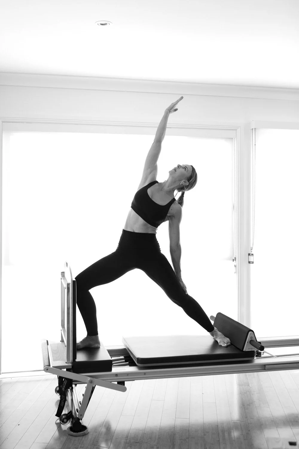 A woman practicing yoga on a Pilates reformer machine indoors, standing in a wide-legged stance with one arm reaching upward and the other hand on her thigh, wearing a sports bra and leggings.