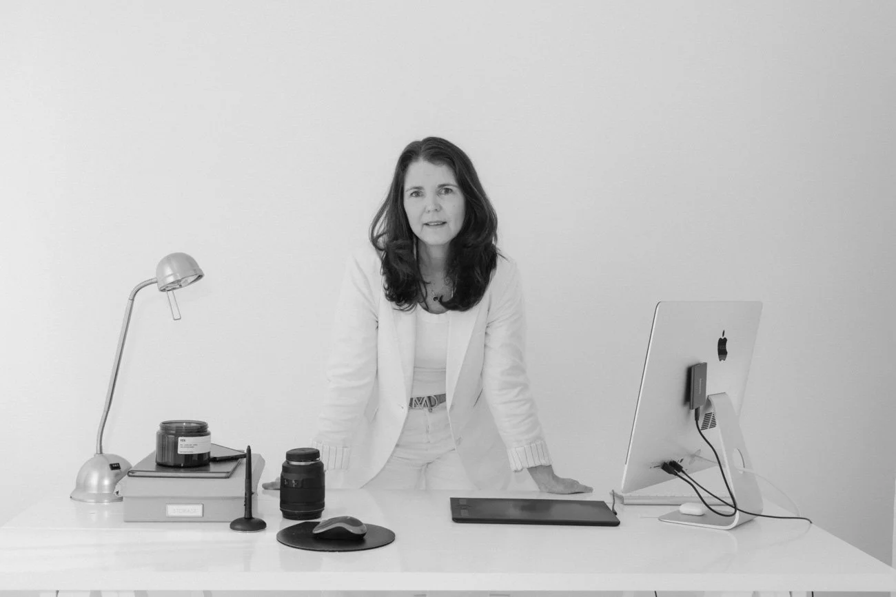 Black and white photo of a woman in a white blazer standing behind a desk with a laptop, camera lens, desk lamp, and office supplies in an office or workspace environment.