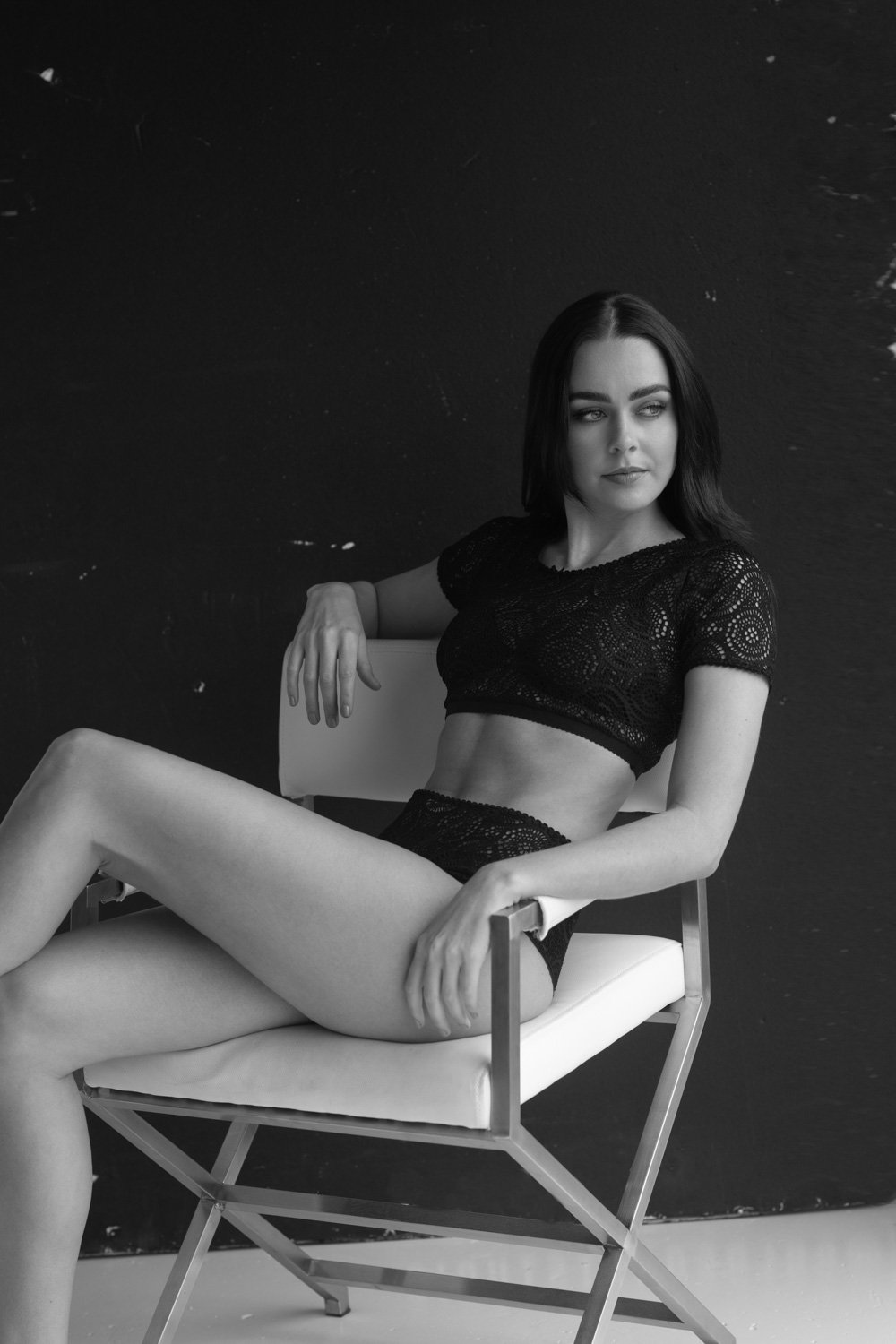 A woman with black hair wearing a black lace crop top and matching underwear sits on a modern white chair against a dark background.