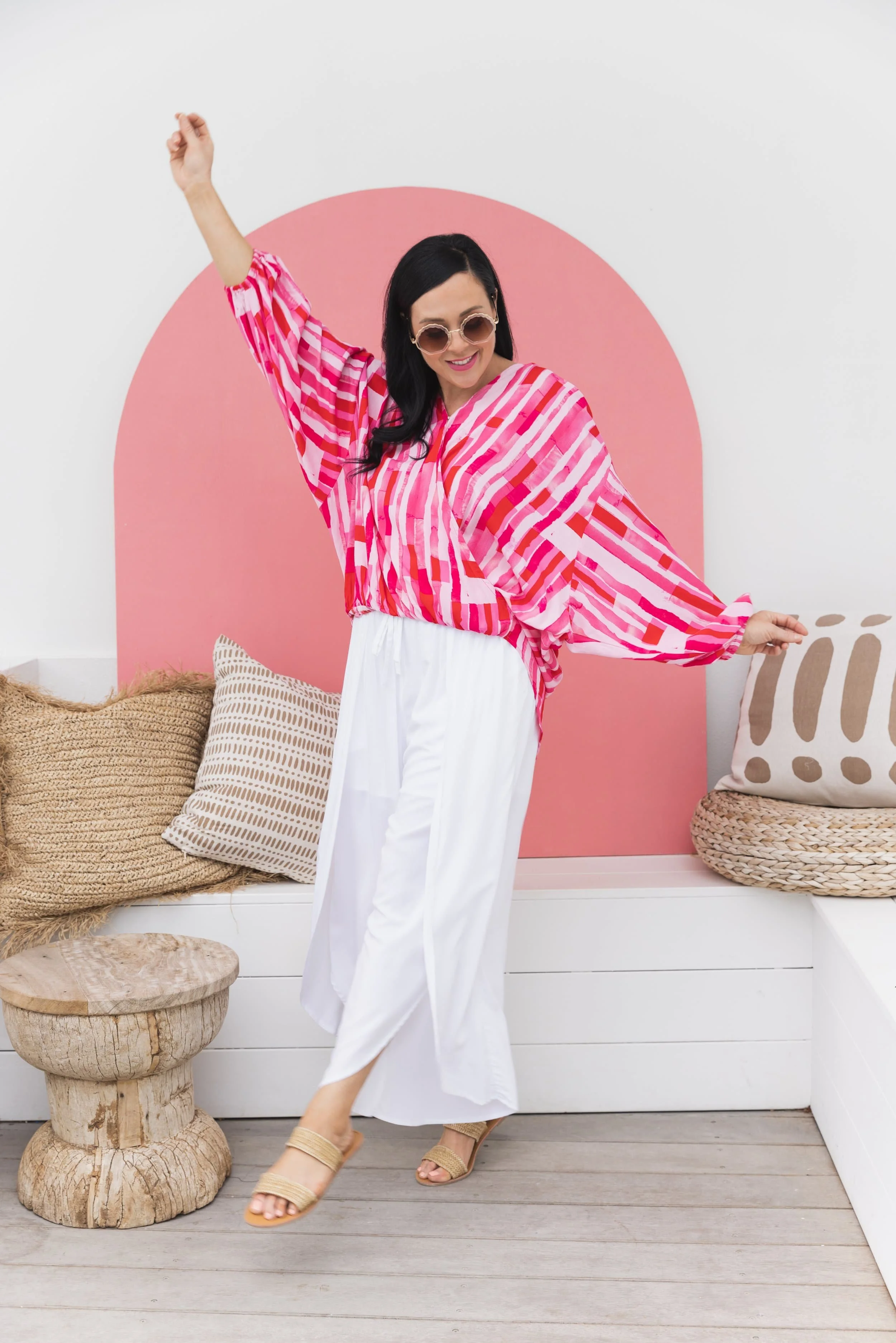 Woman in pink and red patterned blouse and white pants joyful, dancing indoors with a pink backdrop, beige cushions, wooden stool, and wicker basket.