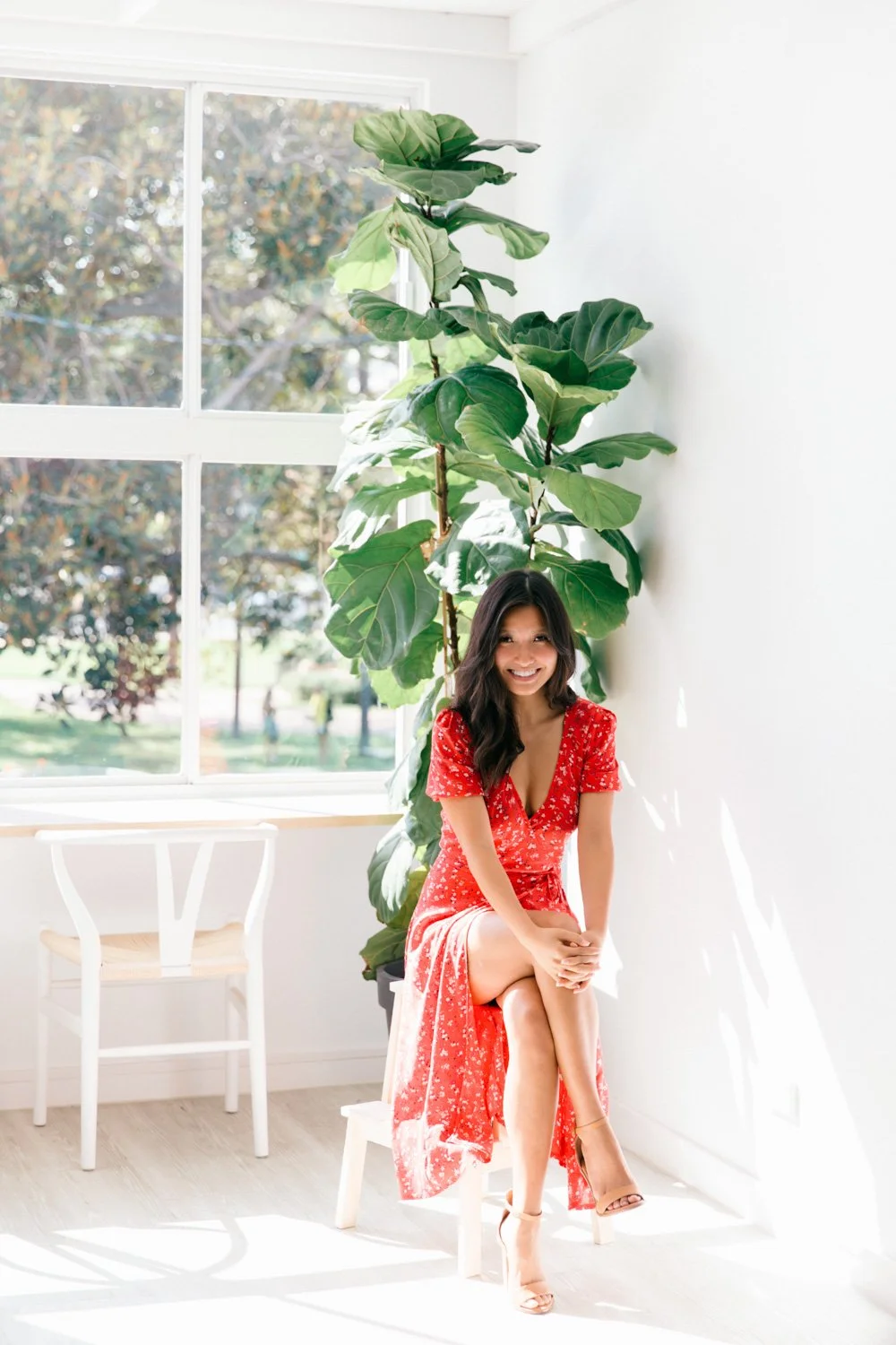 A woman in a red floral dress sitting on a small white chair in a bright room with large windows and a green plant behind her.