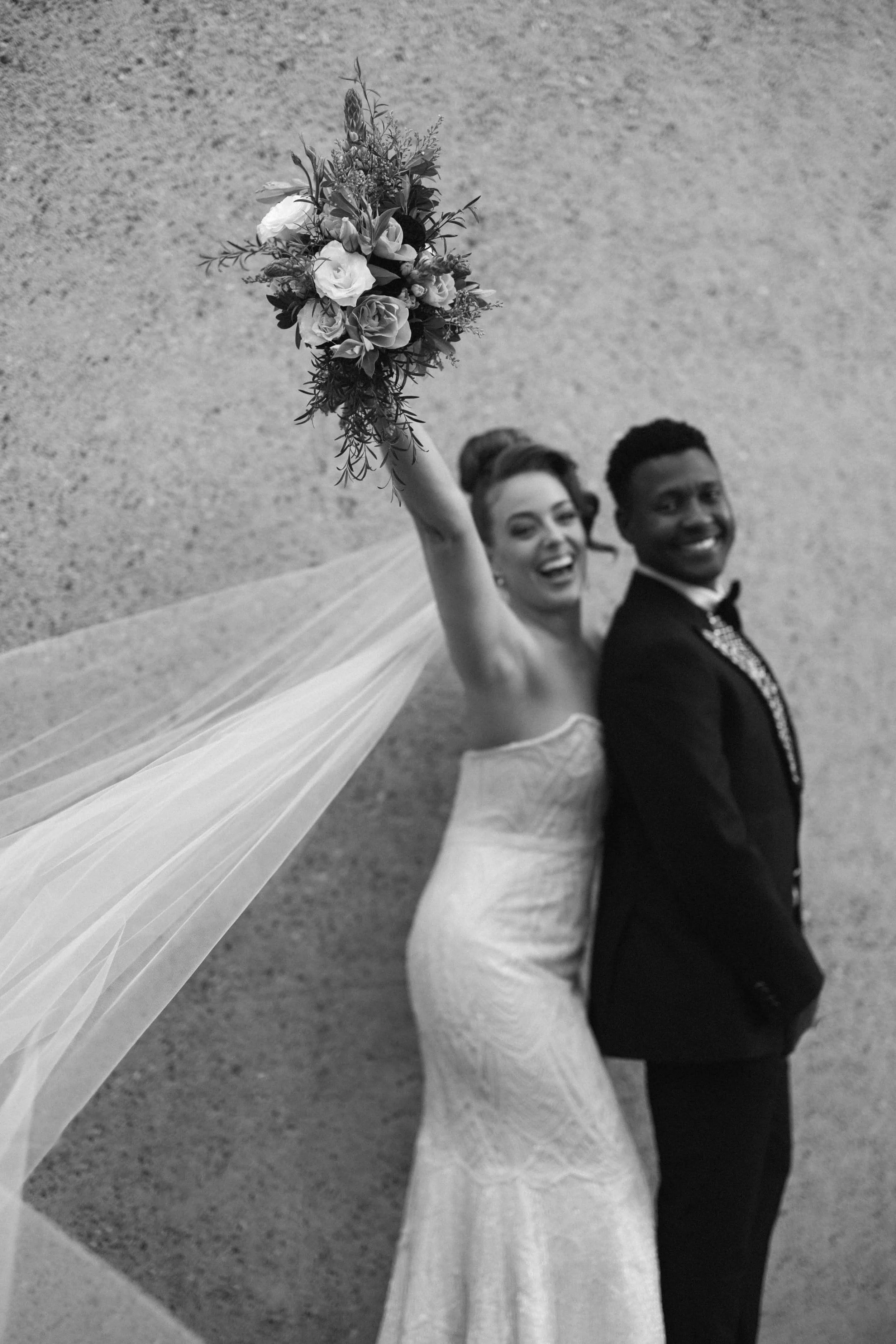 A black-and-white photo of a bride and groom standing back to back, smiling. The bride is holding a bouquet of flowers raised in the air, wearing a strapless wedding gown and a long veil. The groom is wearing a tuxedo with a bow tie. The background a