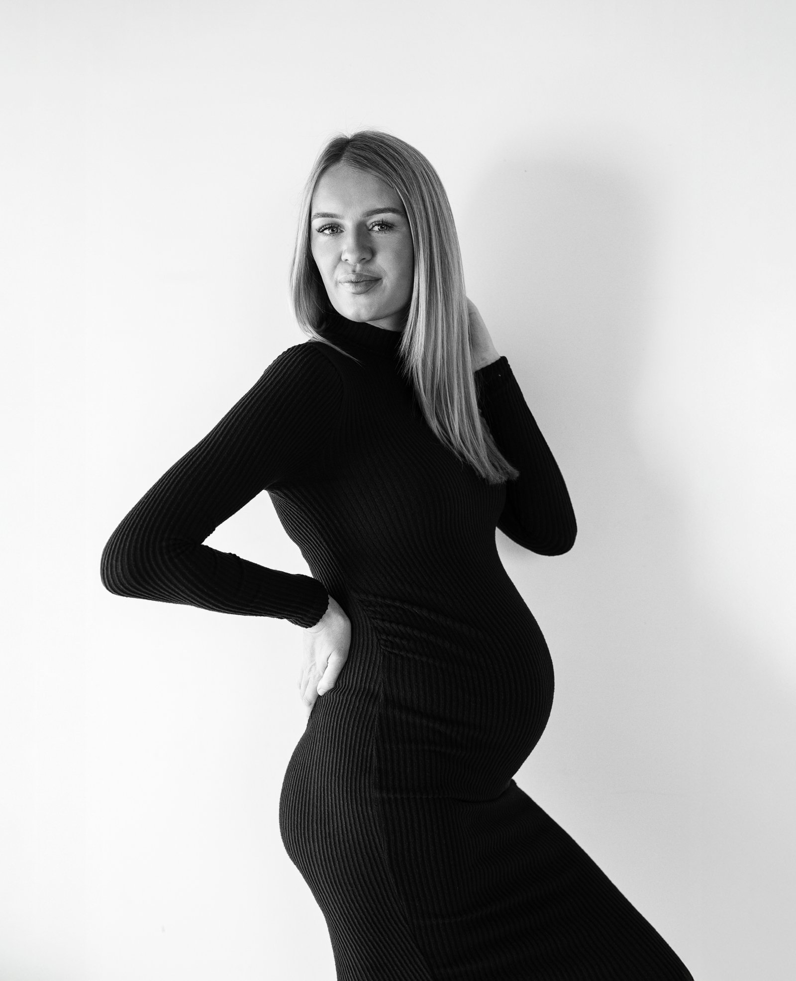 Black and white photo of a pregnant woman wearing a long-sleeved, ribbed, form-fitting dress posing with one hand on her hip and the other touching her neck, standing against a plain wall. Gold Coast maternity photography