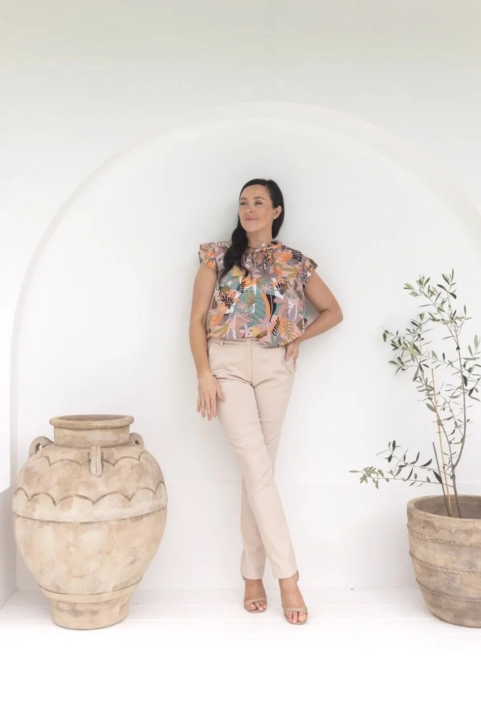 A woman standing against a white wall, wearing a floral top and beige pants, with a large pottery vase and a potted plant beside her.