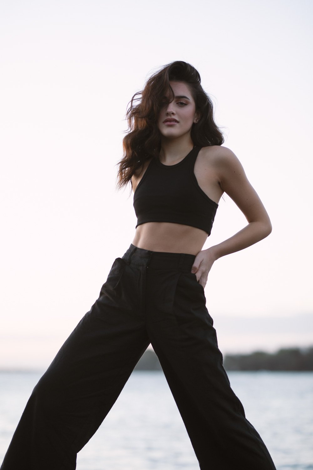 A young woman with long, wavy brown hair standing outdoors near water, wearing a black crop top and high-waisted black pants, posing confidently with one hand on her hip.